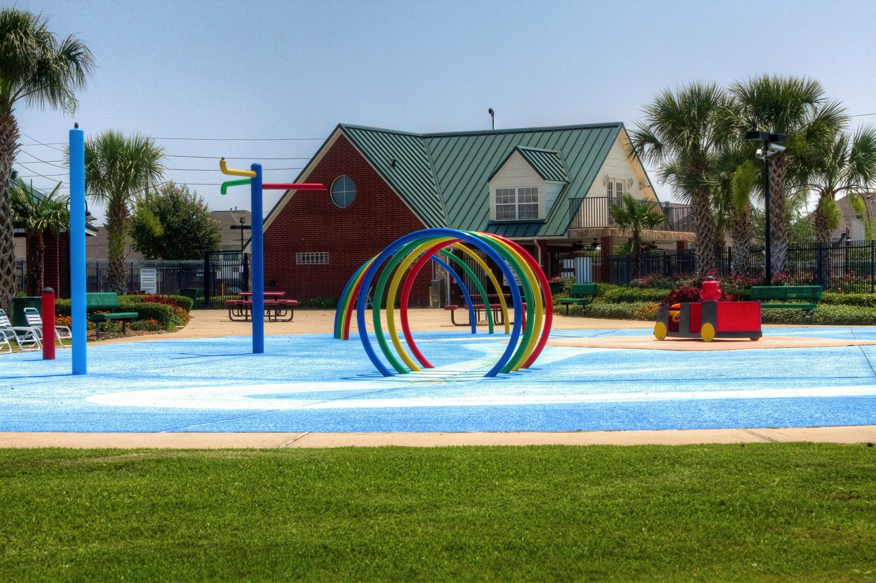 16434 Redcrest Drive Houston, TX 77095 - Photo 40 of 44 This photo features a vibrant community splash pad with colorful play structures, surrounded by lush greenery and palm trees, ideal for family-friendly outdoor activities.