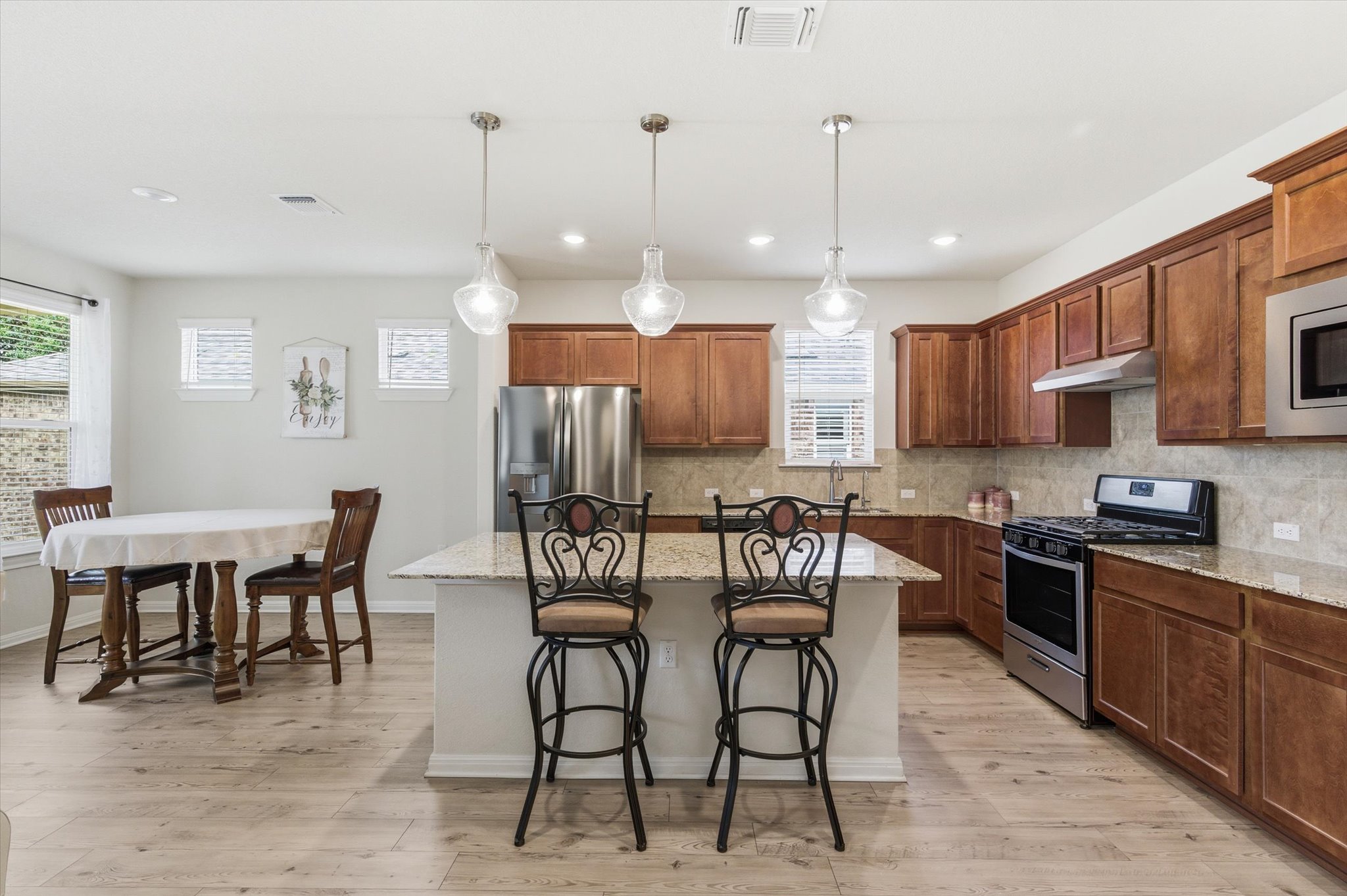 113 Lafayette Street Georgetown, TX 78633 - Photo 10 of 39 Open-concept kitchen and dining area featuring light wood-finish flooring, a kitchen island with pendant lighting, stainless steel appliances, abundant cabinetry, and granite countertops with a coordinating backsplash