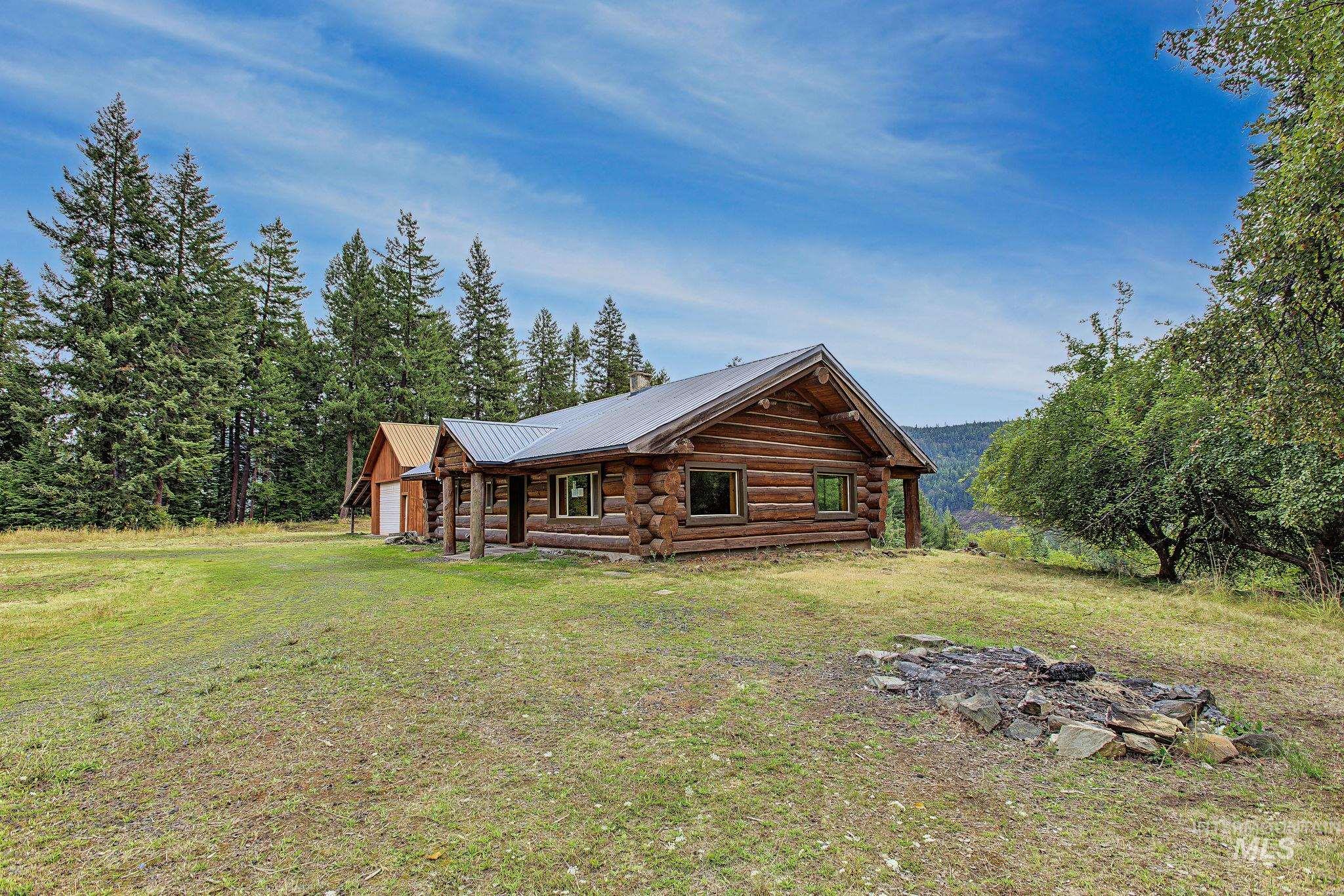 2926 Wells Bench Road Orofino, ID 83544 - Photo 44 of 48 Log cabin exterior with a metal roof, surrounded by mature trees, a detached outbuilding, and a stone-lined fire pit