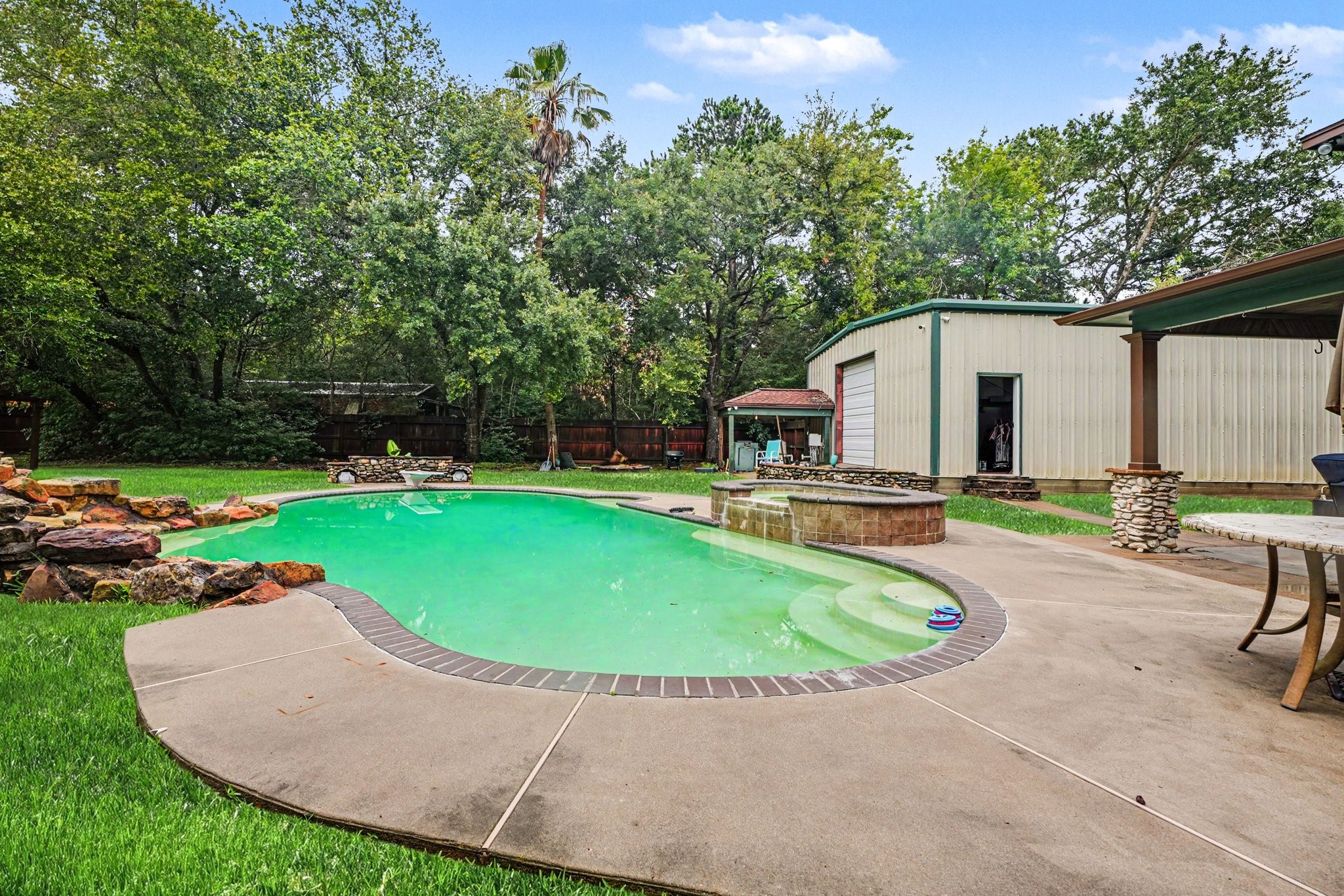 5104 Avenue R Santa Fe, TX 77510 - Photo 15 of 35 This backyard features a spacious pool with a hot tub, surrounded by a concrete patio and lush greenery. There's a large workshop and a covered seating area, perfect for outdoor entertaining.