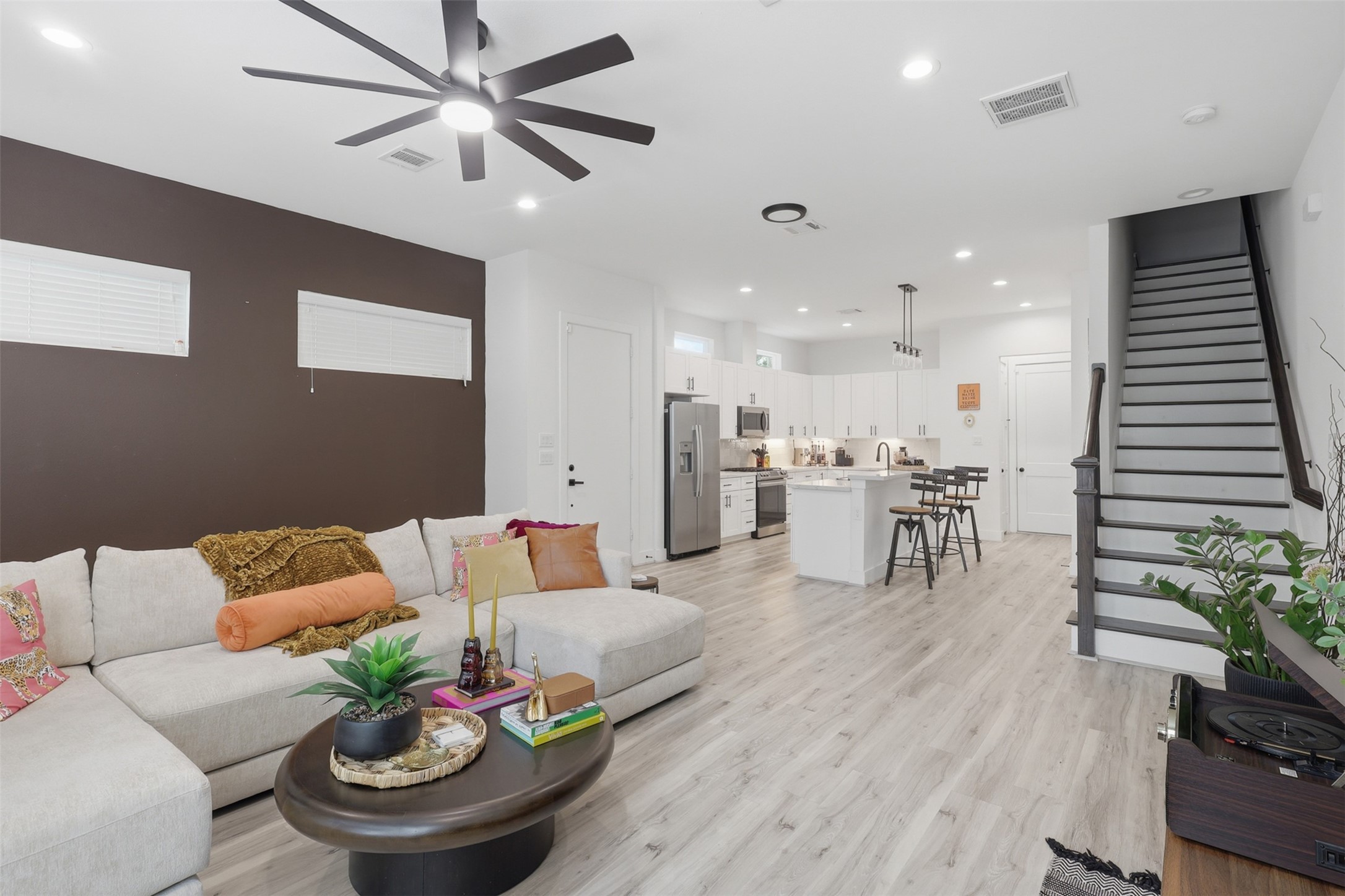 6833 Paris Street Houston, TX 77021 - Photo 2 of 23 This modern living space features an open-concept layout with a cozy sectional, sleek ceiling fan, and a dining area. The bright kitchen includes stainless steel appliances and an island with bar seating. A staircase leads to the upper level, and natural light enhances the inviting atmosphere.