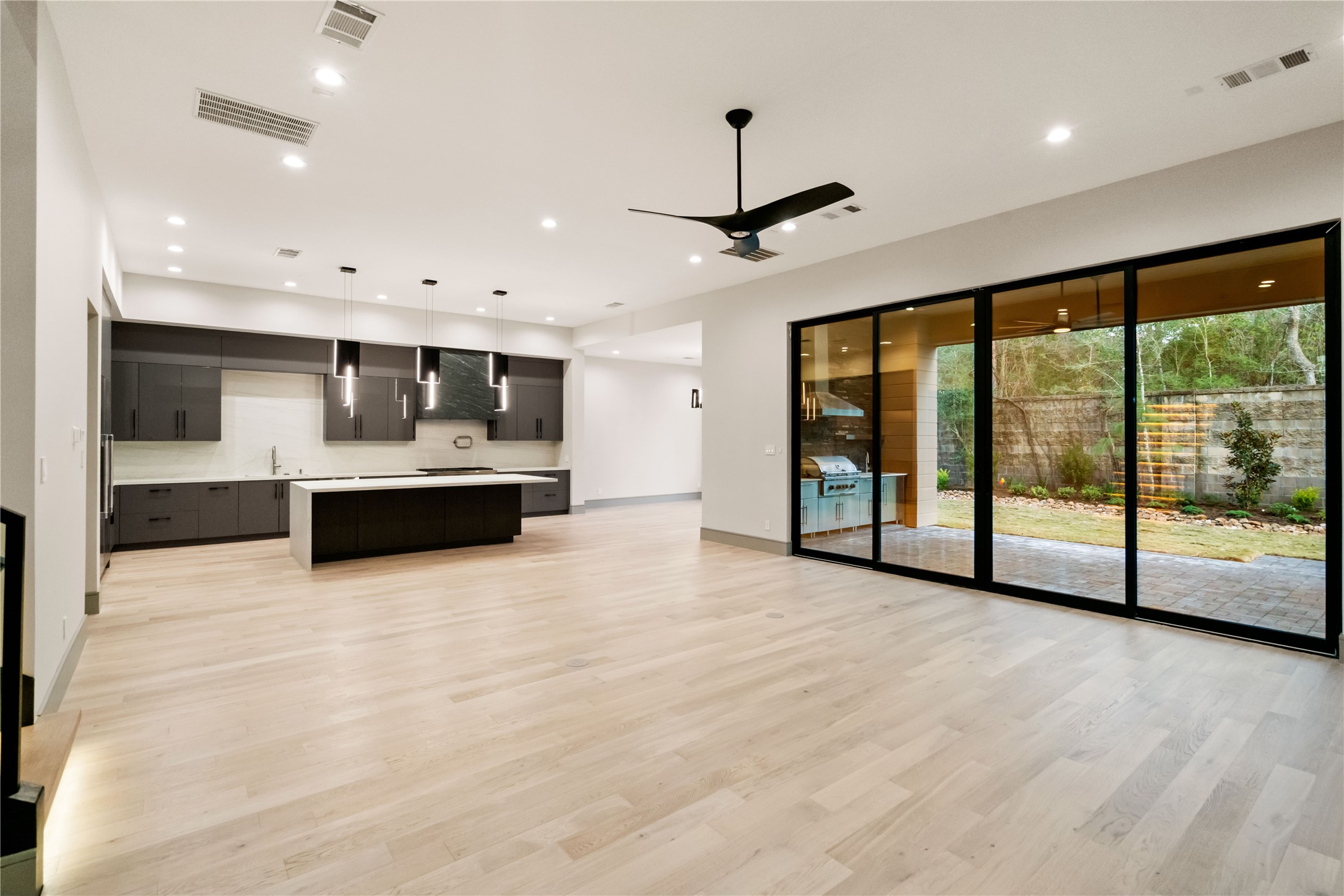 47 Pronghorn Place Spring, TX 77389 - Photo 6 of 44 This modern open-concept living space features a sleek kitchen with dark cabinetry, a large island, and pendant lighting. It has light wood flooring and expansive sliding glass doors leading to a patio and landscaped yard, offering ample natural light and outdoor views.
