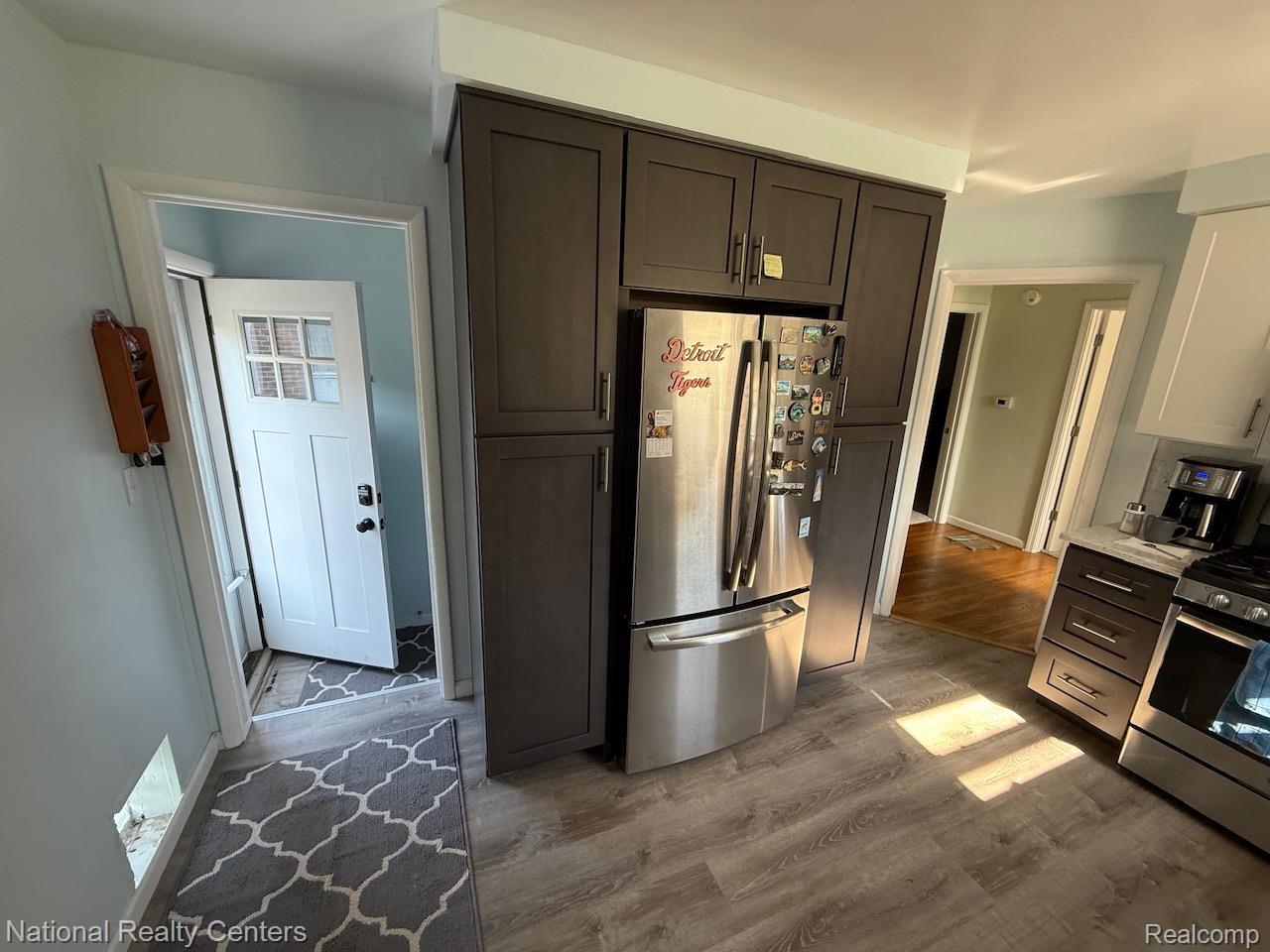 Full-height cabinetry, stainless steel refrigerator, white shaker-style upper cabinets, wood-finish flooring, and white paneled door with glass inserts