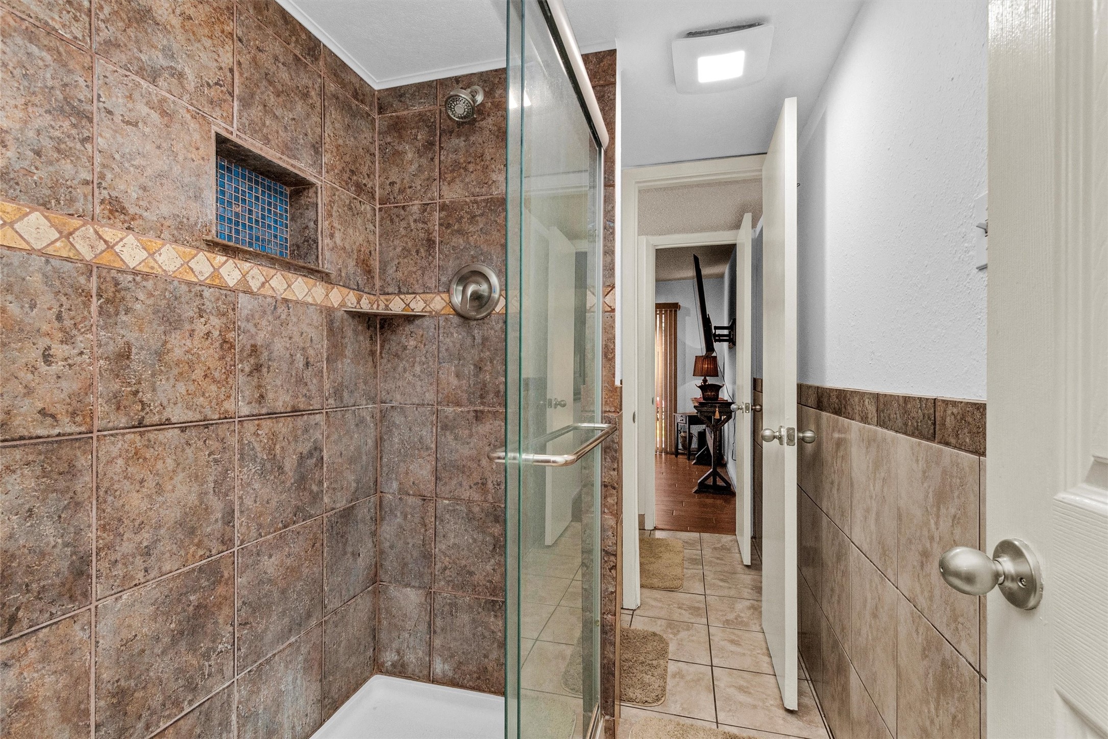 12900 Walden Road, Unit D 411 Montgomery, TX 77356 - Photo 11 of 18 Discover elegance in this bathroom's tiled shower with a chic glass door, featuring a decorative tile strip and niche, all leading into a beautifully tiled hallway that flows seamlessly into a room with warm wood flooring.