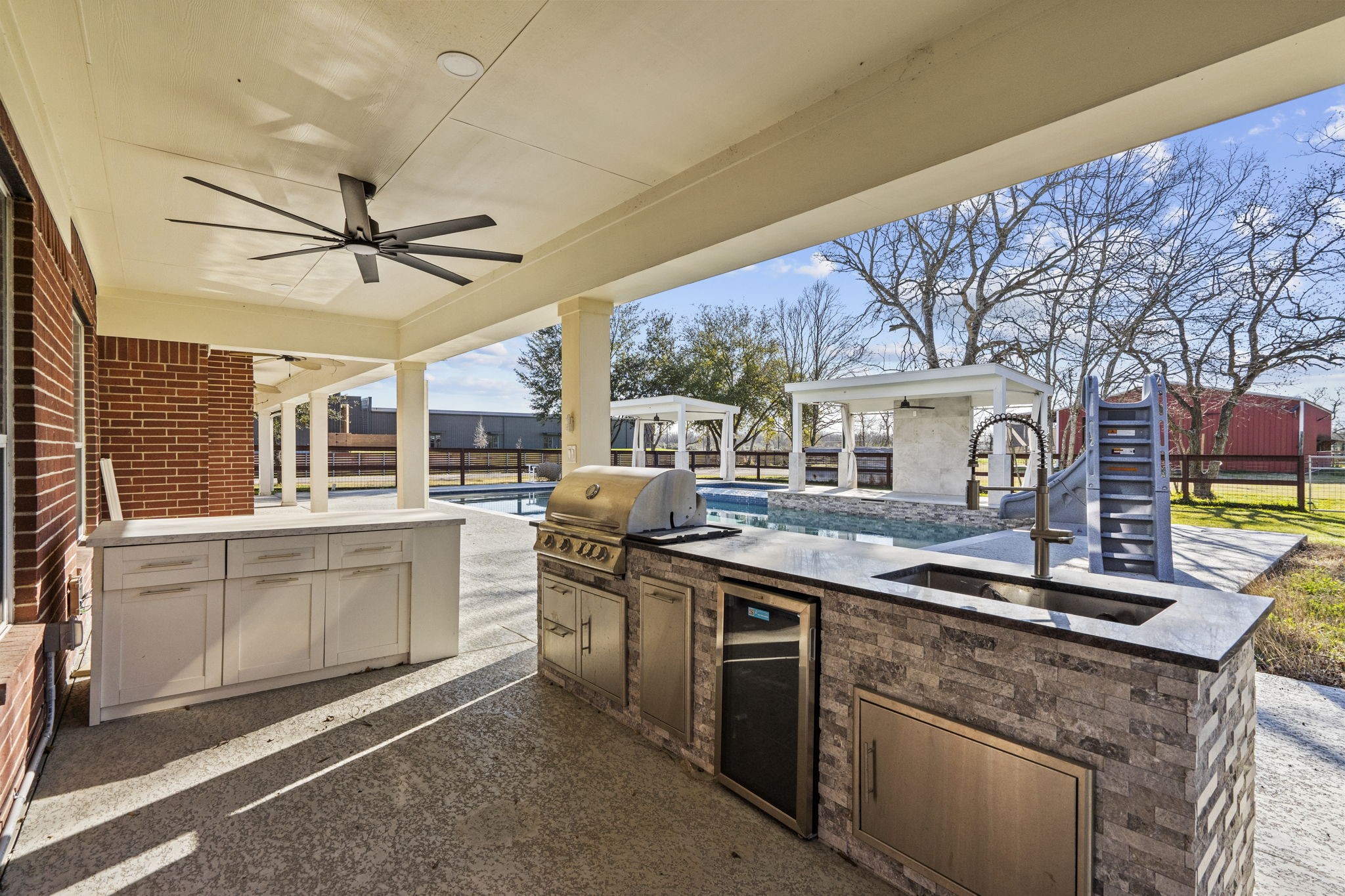2730 Lawrence Road Kemah, TX 77565 - Photo 42 of 46 This photo showcases a spacious outdoor kitchen and entertainment area featuring a built-in grill, sink, and ample counter space. Adjacent is a swimming pool with a slide, surrounded by a fenced yard and shaded by mature trees, perfect for hosting gatherings.