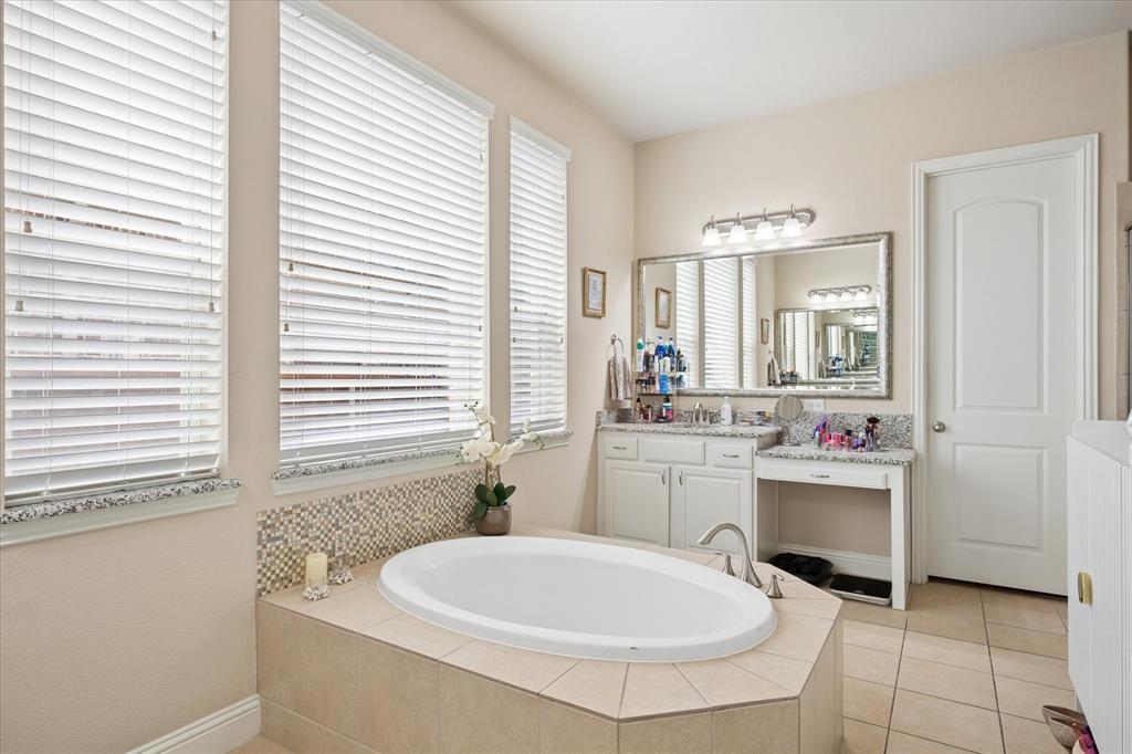 8268 Kara Creek Road Frisco, TX 75036 - Photo 13 of 31 Bathroom featuring a built-in soaking tub with mosaic tile surround, multiple windows with blinds, a dual-sink vanity with a large framed mirror, and a separate vanity with a granite-style countertop