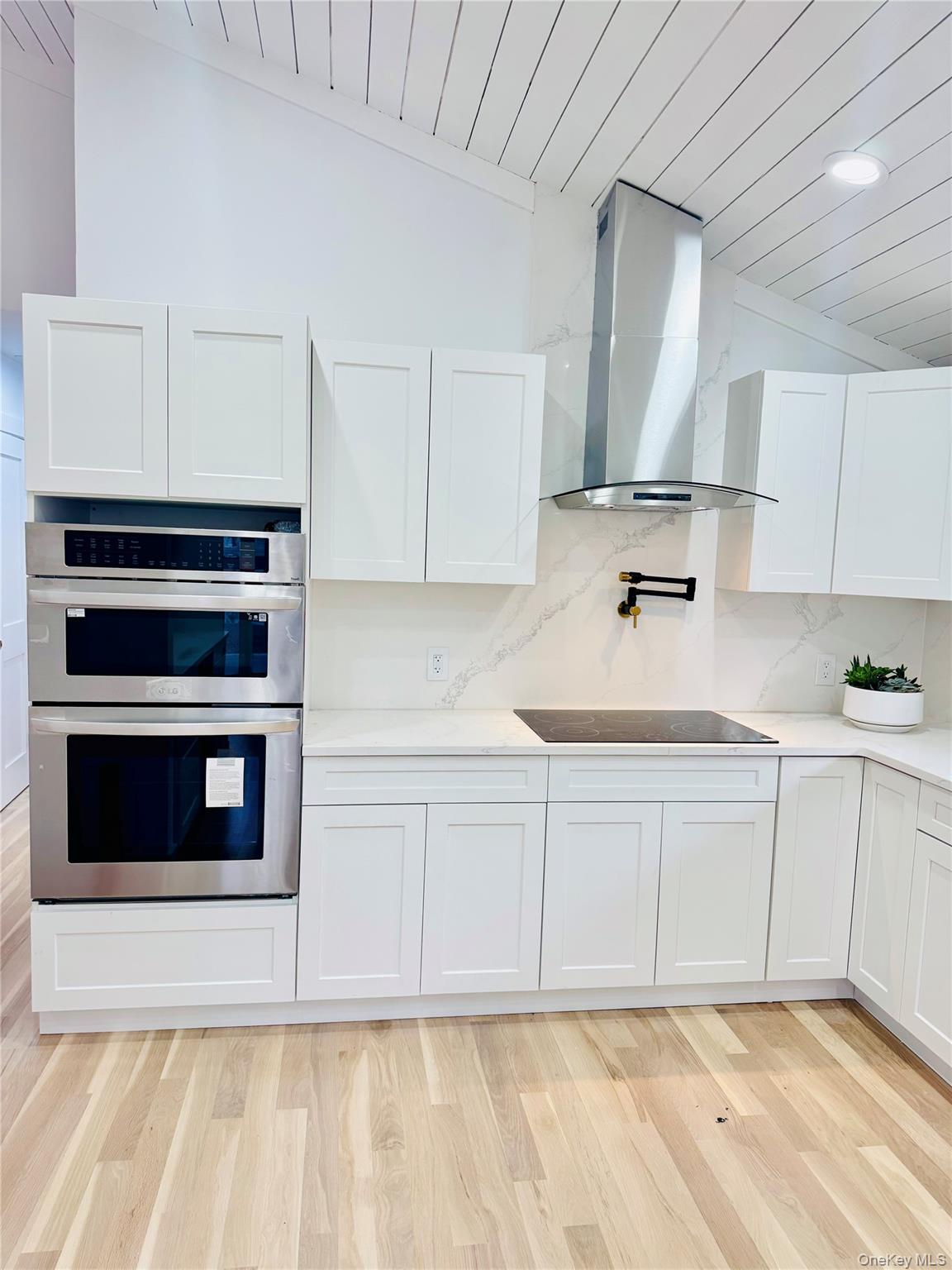 105 Old Riverhead Road Hampton Bays, NY 11946 - Photo 4 of 18 Kitchen featuring stainless steel double oven, white cabinetry, light wood-style flooring, wall chimney exhaust hood, and decorative backsplash