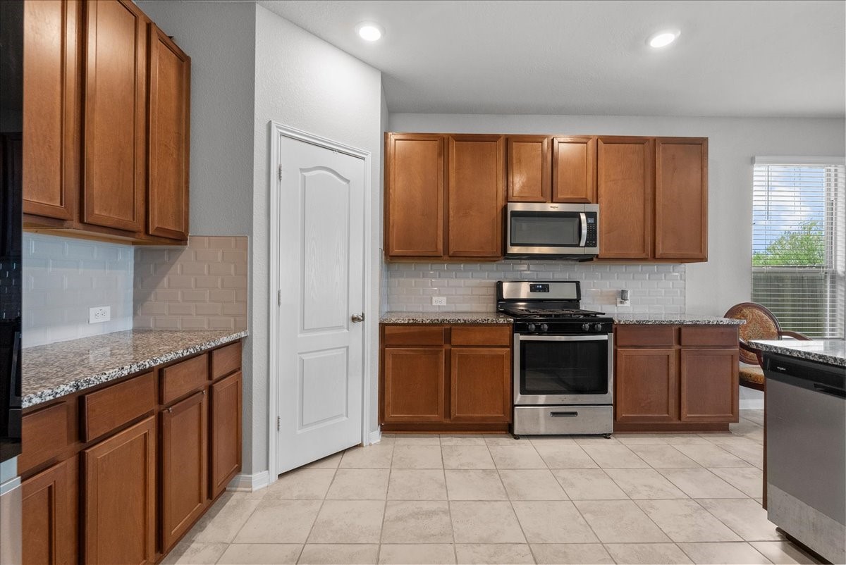 2320 Rosemary Street Rosenberg, TX 77469 - Photo 13 of 40 This kitchen features modern stainless steel appliances, ample wooden cabinetry, and granite countertops. It has a sleek, neutral tile floor and stylish white subway tile backsplash, creating a bright and inviting space.