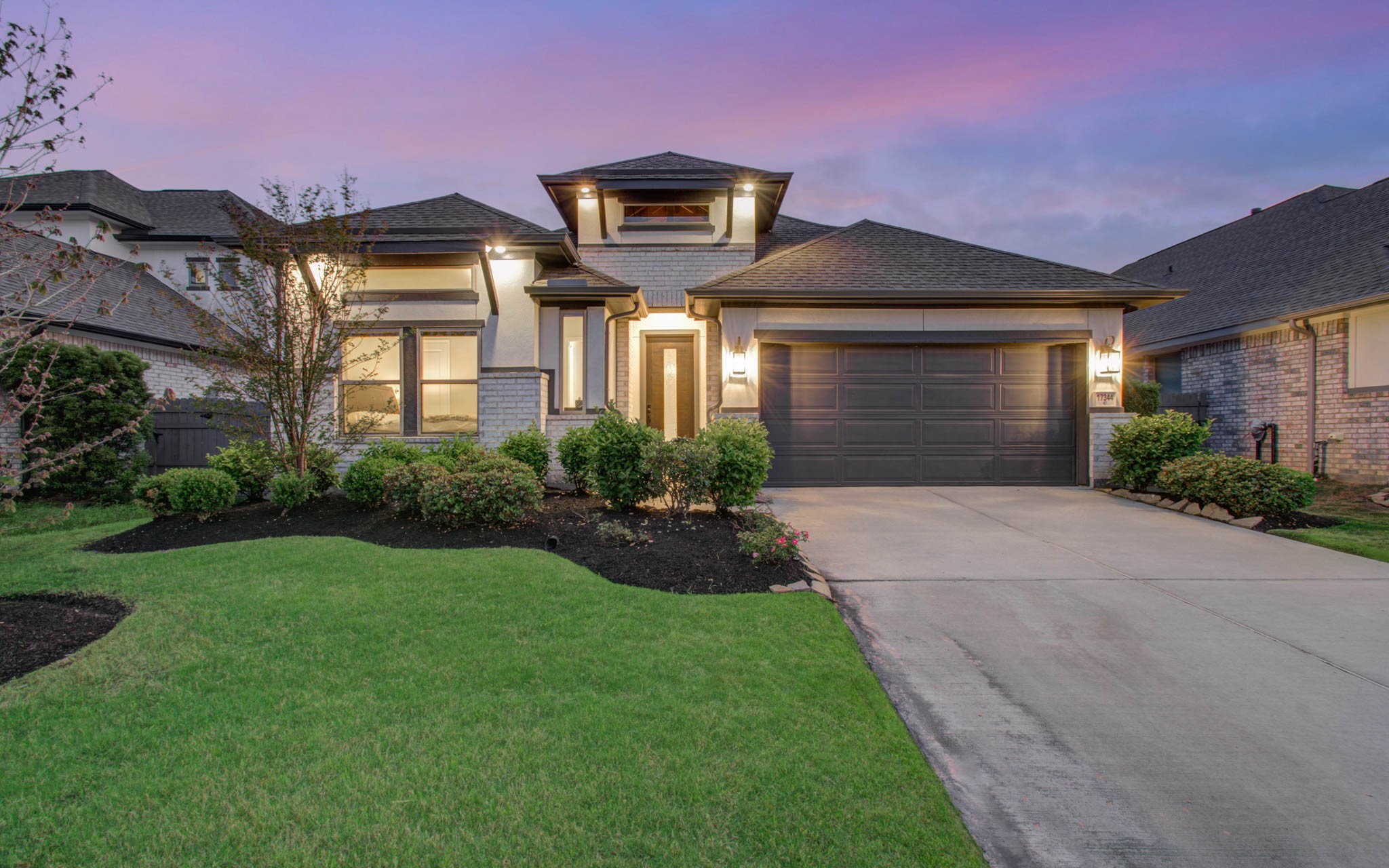 17344 Grand Canyon Road Conroe, TX 77302 - Photo 41 of 49 This photo showcases a modern, single-story home with a two-car garage and a well-maintained front yard. The exterior features a combination of stone and stucco, with warm exterior lighting highlighting the entrance. The driveway is spacious, and the landscaping includes shrubs and a neat lawn, all set against a vibrant sunset sky.