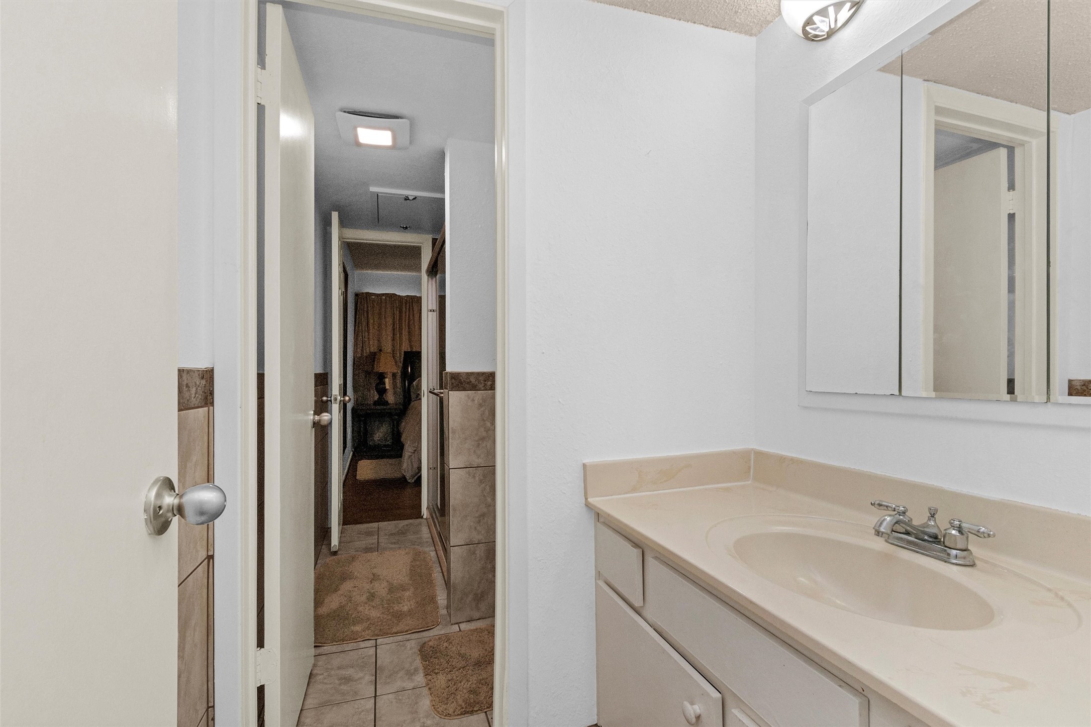12900 Walden Road, Unit D 411 Montgomery, TX 77356 - Photo 9 of 18 This inviting bathroom features a sleek single sink vanity, a spacious mirror, and light-colored tile flooring, seamlessly leading into a cozy carpeted hallway with a glimpse of another room beyond.
