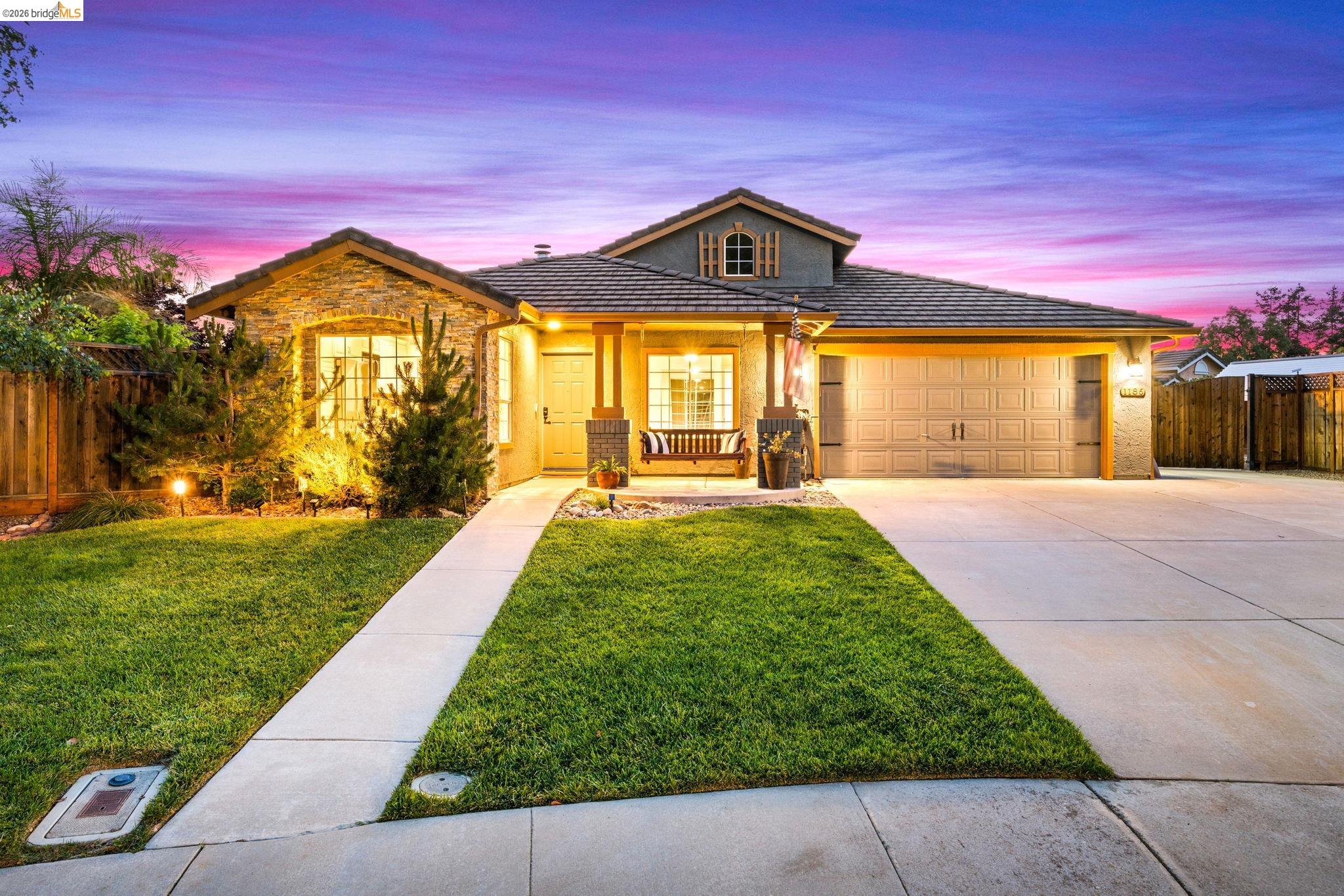 1186 El Rio Court Livermore, CA 94551 - Photo 4 of 60 Stone and stucco exterior featuring an arched window, a covered entryway with a porch swing, a two-car garage, and a concrete driveway