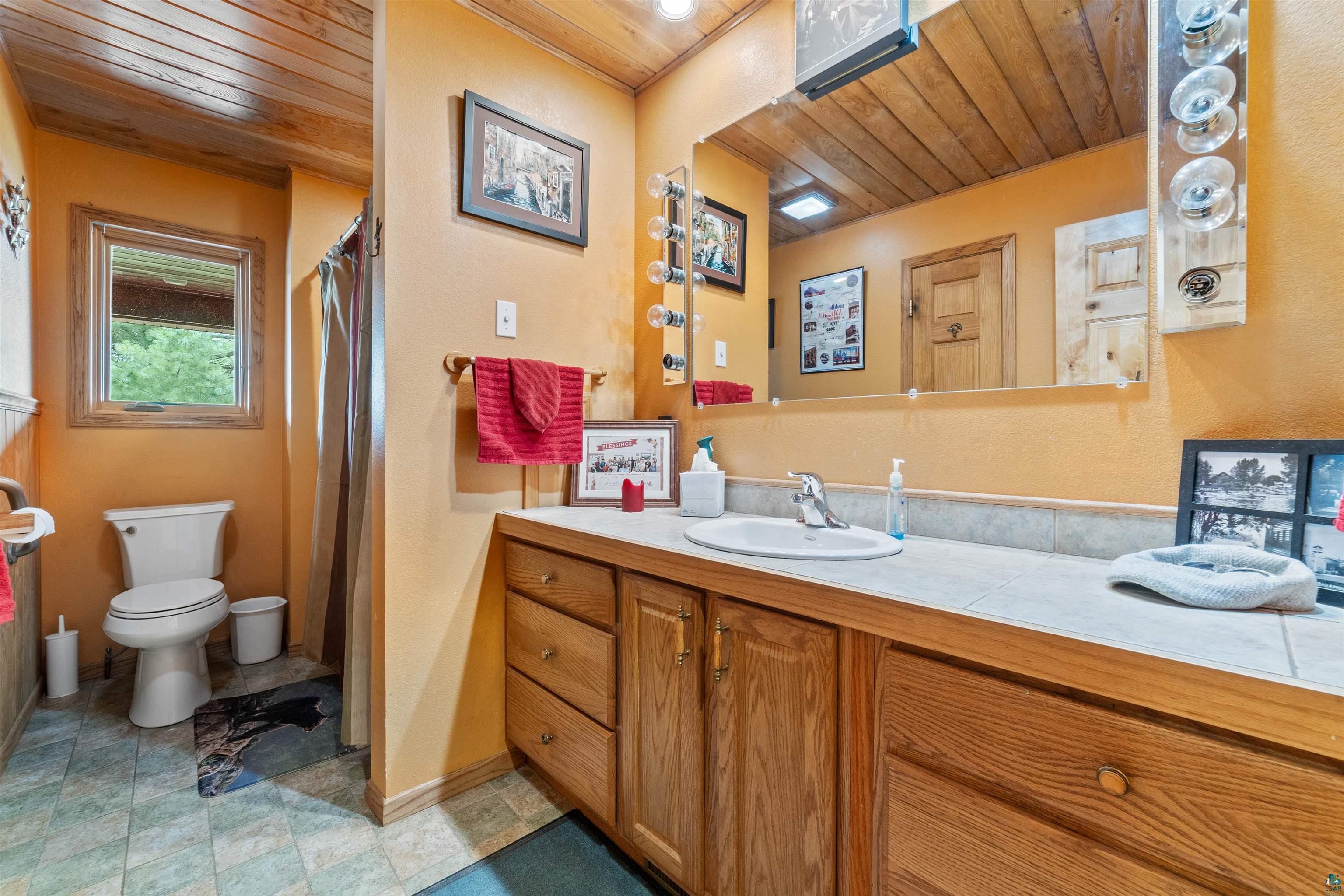 11911 East Rowe Road Lake Nebagamon, WI 54849 - Photo 46 of 61 Bathroom featuring a wood-panel ceiling, spacious vanity with integrated sink, tile countertop, wood-finish cabinetry, and a window