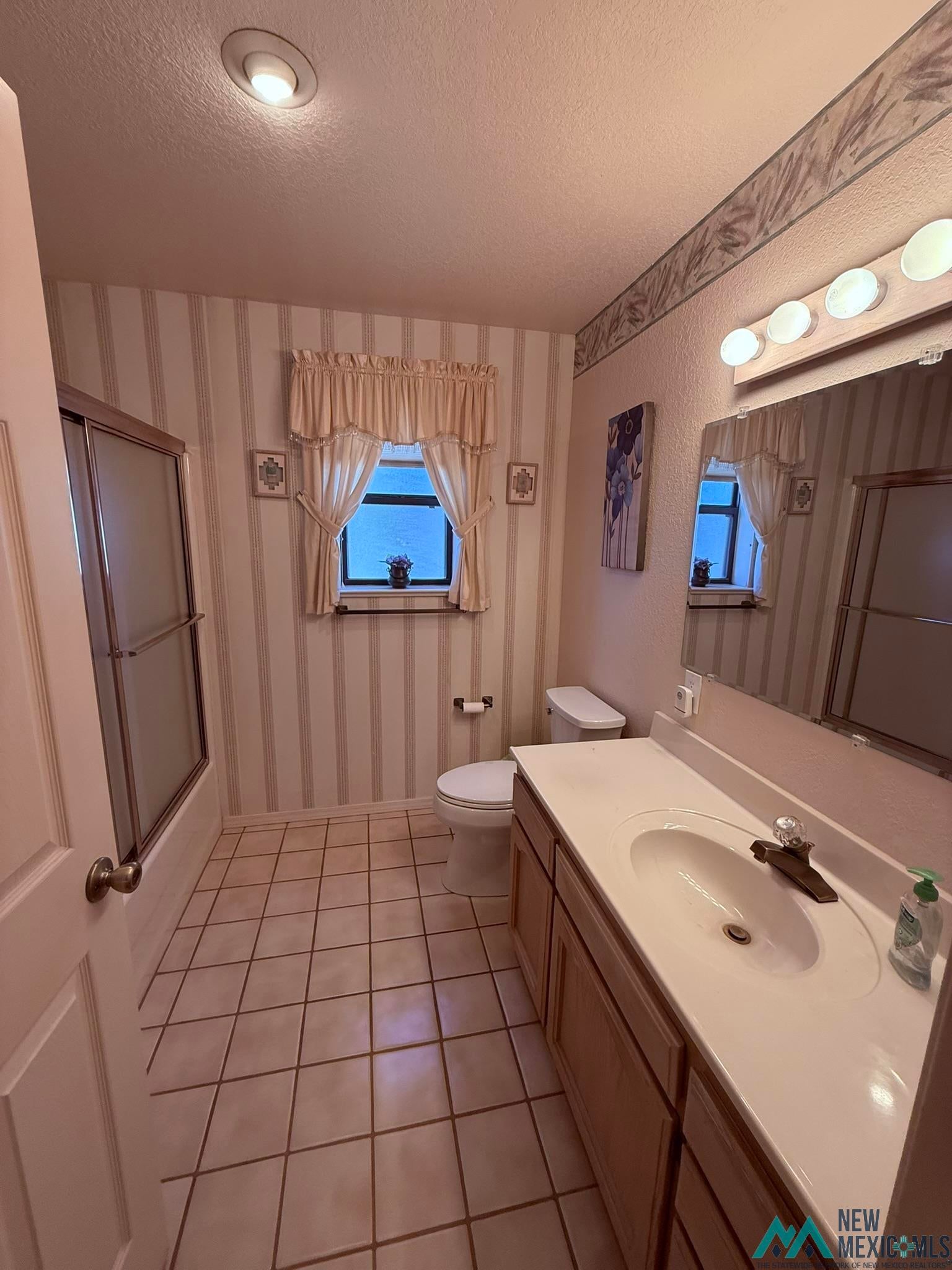 300 Rio Grande Circle Elephant Butte, NM 87935 - Photo 9 of 99 Bathroom featuring a single vanity with integrated sink, a rectangular mirror with strip lighting, a window with valance and curtains, a toilet, and a bathtub with sliding glass doors