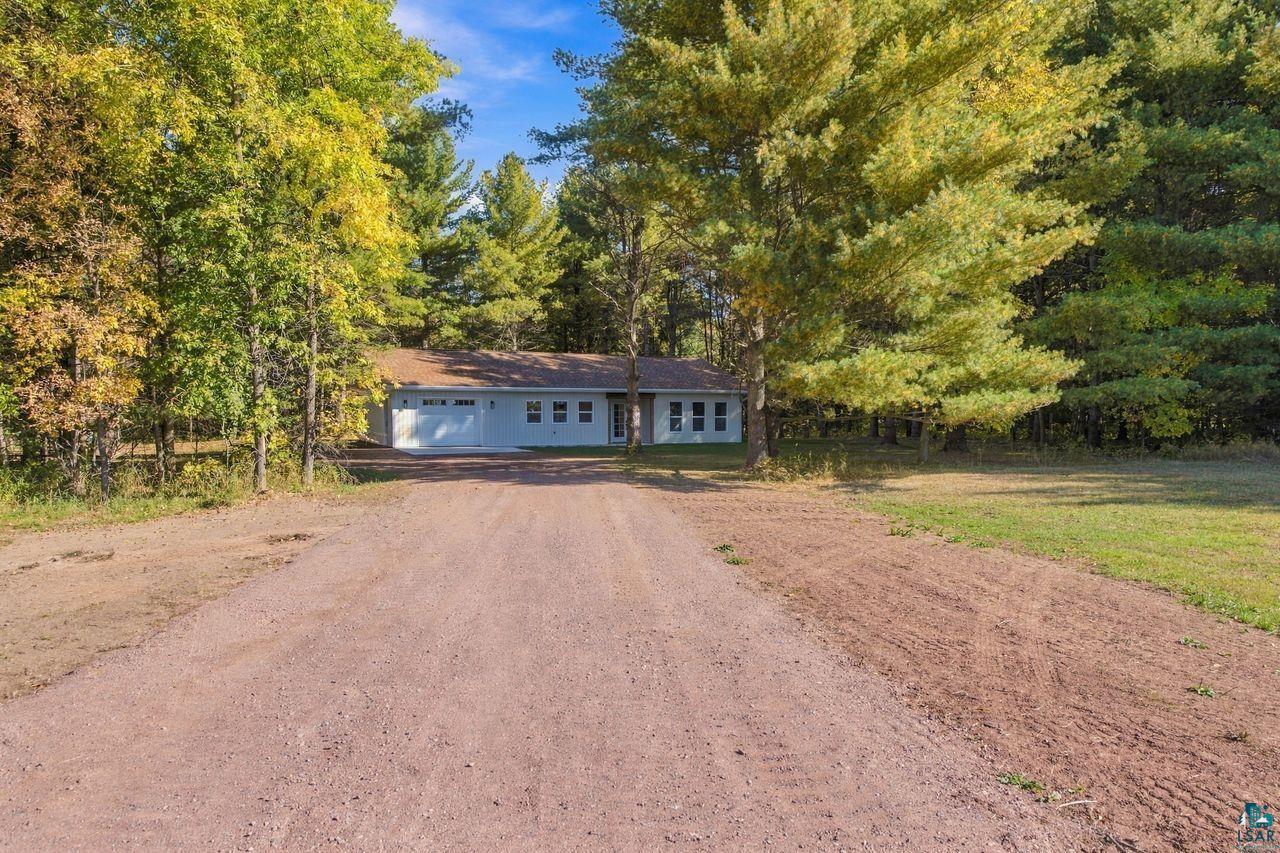 65352 Adler Road Marengo, WI 54855 - Photo 2 of 24 Single-story residence surrounded by mature trees, featuring a light-colored exterior and a two-car garage