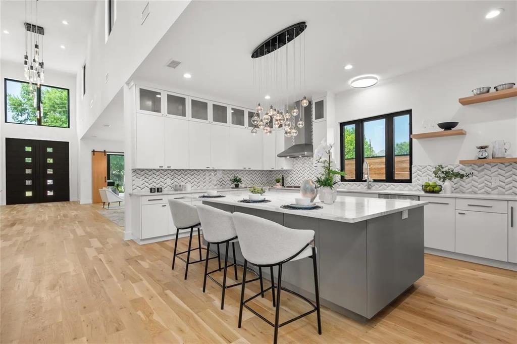 309 Kaye Street Coppell, TX 75019 - Photo 31 of 36 Open-concept kitchen with a large central island, light wood-finish flooring, white cabinetry with glass upper doors, a chevron pattern tile backsplash, and contemporary pendant lighting