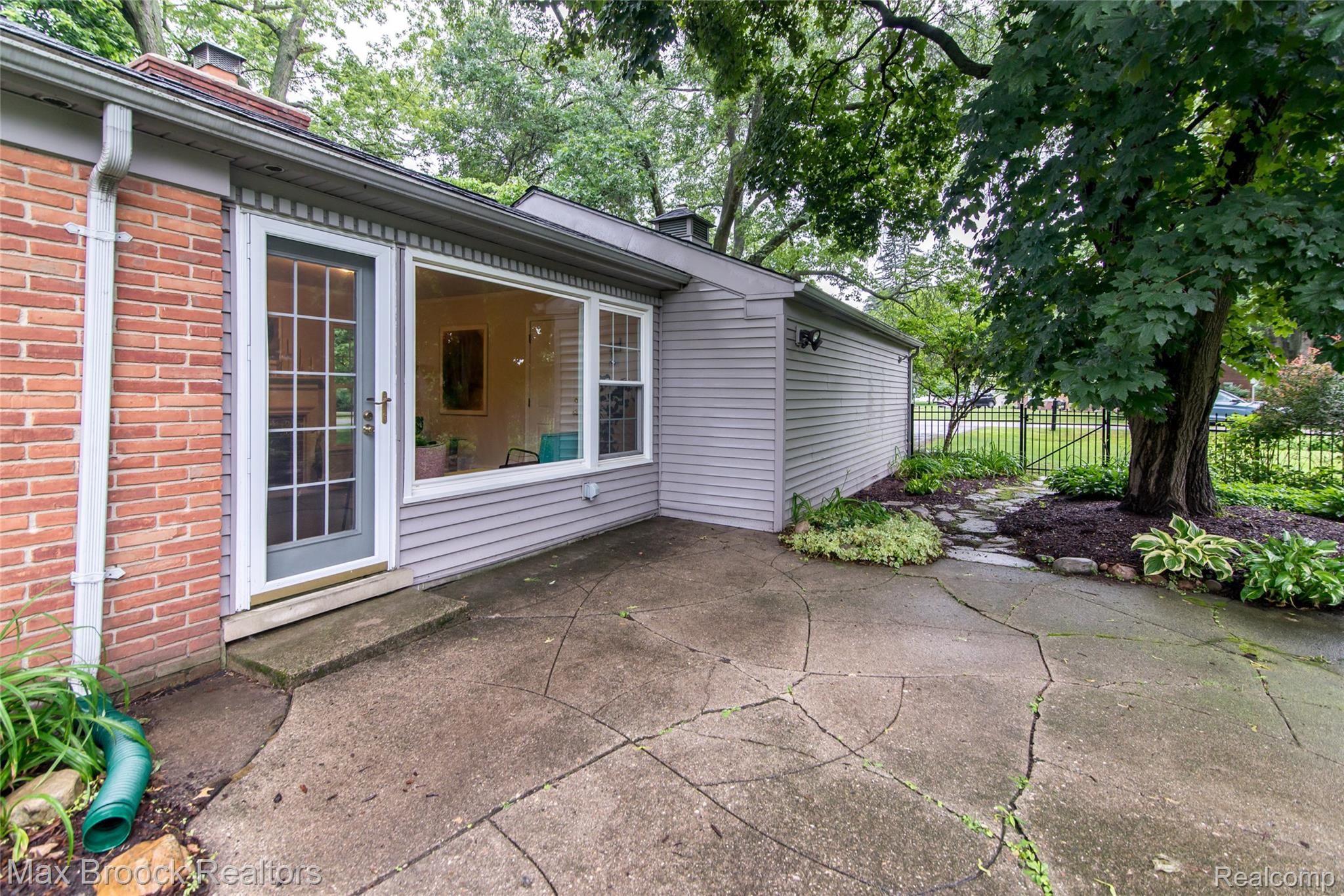 6799 East Dartmoor Road West Bloomfield, MI 48322 - Photo 26 of 36 Concrete patio with a full-light paneled door, large picture window, brick and siding exterior, and an adjacent landscaped area with mature trees and a stone pathway