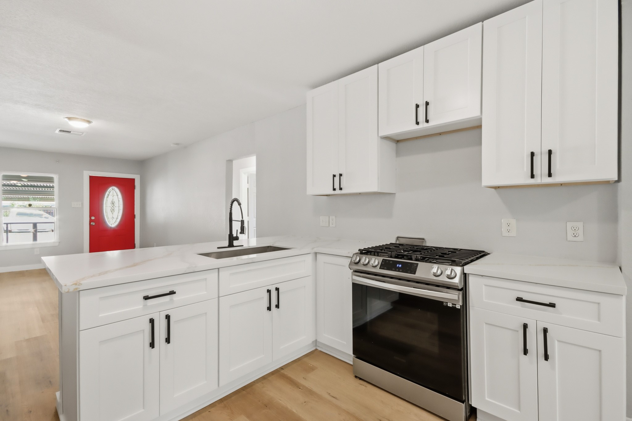 10430 Wiggins Street Houston, TX 77029 - Photo 13 of 27 This kitchen features modern white cabinetry, a sleek black faucet, and stainless steel appliances, including a gas stove. The open layout flows into a bright living area with a striking red front door.