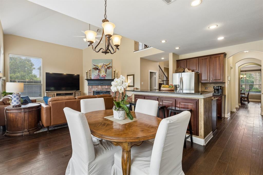 960 Remuda Road Frisco, TX 75033 - Photo 20 of 39 Open-concept living space featuring wood-finish flooring, a kitchen island with dark wood cabinetry and a stone countertop, a fireplace with a brick surround, and a vaulted ceiling with a chandelier