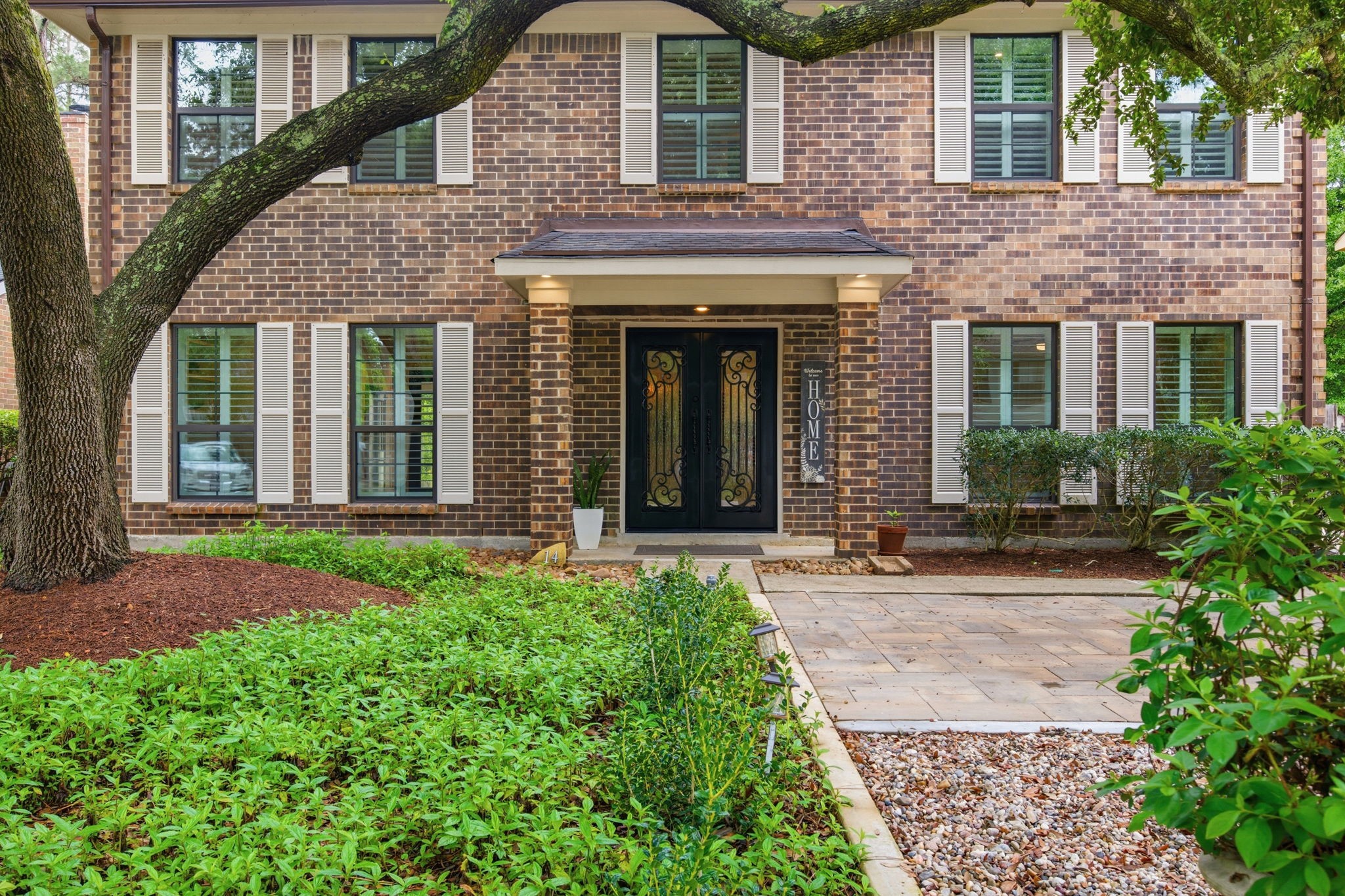 14 Ridgeline Court Spring, TX 77381 - Photo 3 of 40 This home features a welcoming front entrance with elegant iron double doors, surrounded by lush greenery and mature trees. It offers a classic facade with shutters on the upgraded windows and extended tile pavers providing additional parking.