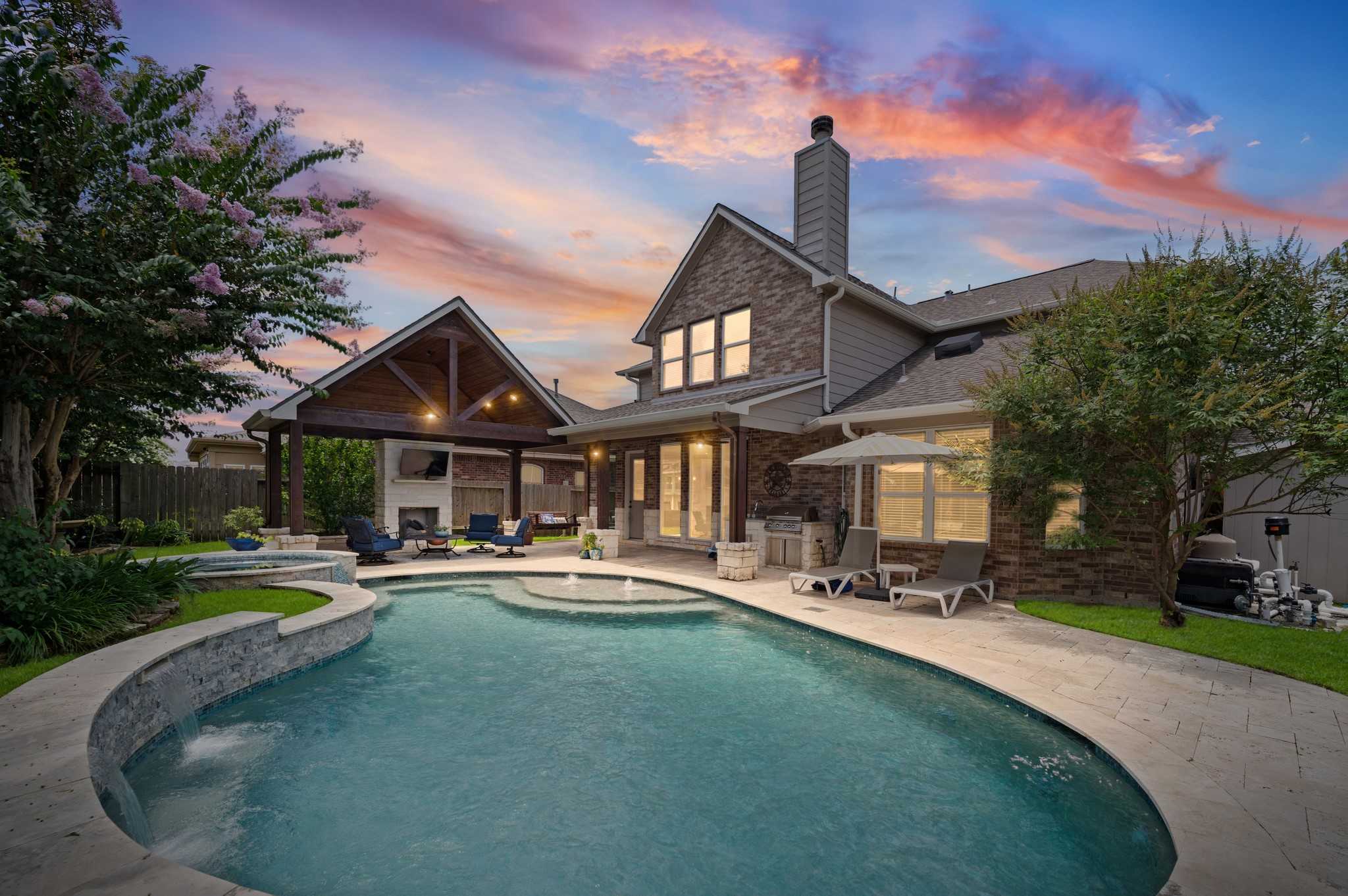 9507 Empress Crossing Drive Spring, TX 77379 - Photo 1 of 50 This stunning home features a beautiful backyard with a curvy pool, spacious patio, and cozy covered seating area with a fireplace. Perfect for entertaining or relaxing, the setting is enhanced by a vibrant sunset sky.