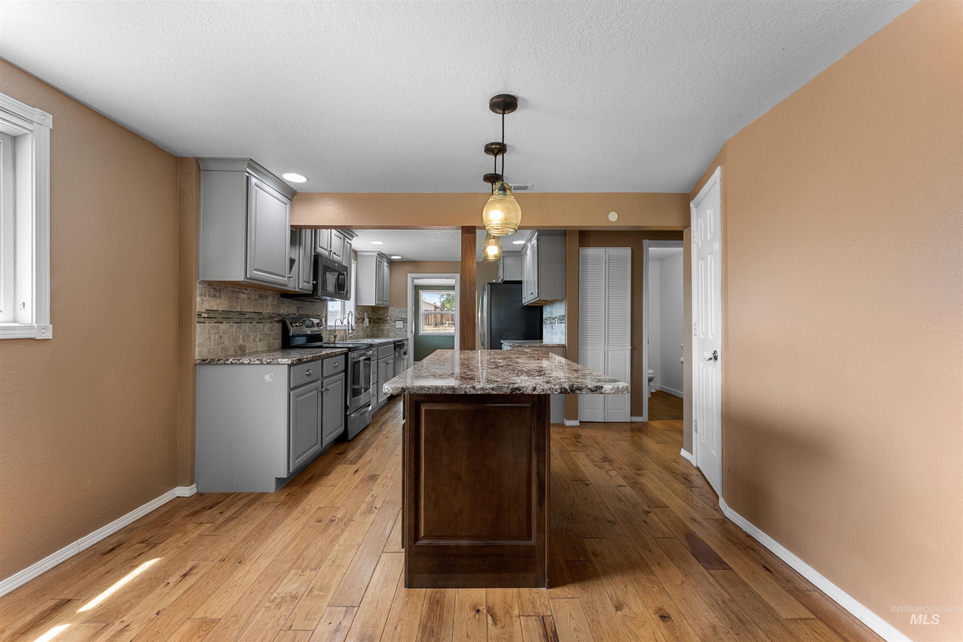 17441 Upper Pleasant Ridge Road Caldwell, ID 83607 - Photo 9 of 40 Kitchen featuring wood-finish flooring, a dark wood island with a stone countertop, grey cabinetry, stainless steel appliances, and an intricate backsplash