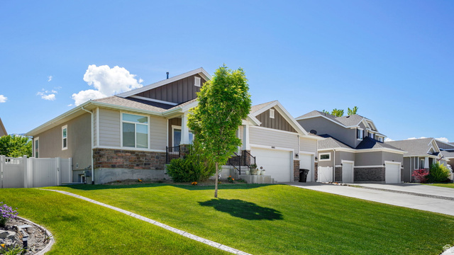 6062 Spring Street Stansbury Park, UT 84074 - Photo 2 of 36 Contemporary home featuring a stone and siding facade, a manicured front lawn, a white picket fence, a dedicated driveway, and an attached two-car garage