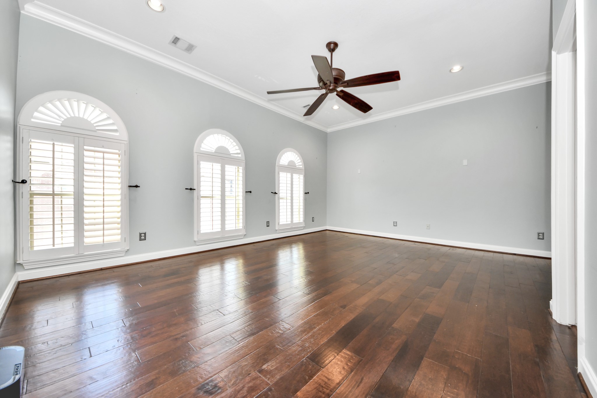 710 Jackson Hill Street Houston, TX 77007 - Photo 22 of 31 Spacious Primary bedroom with hardwood floors, light gray walls, and crown molding. Features three arched windows with shutters, a ceiling fan, and recessed lighting, offering a bright and airy feel.