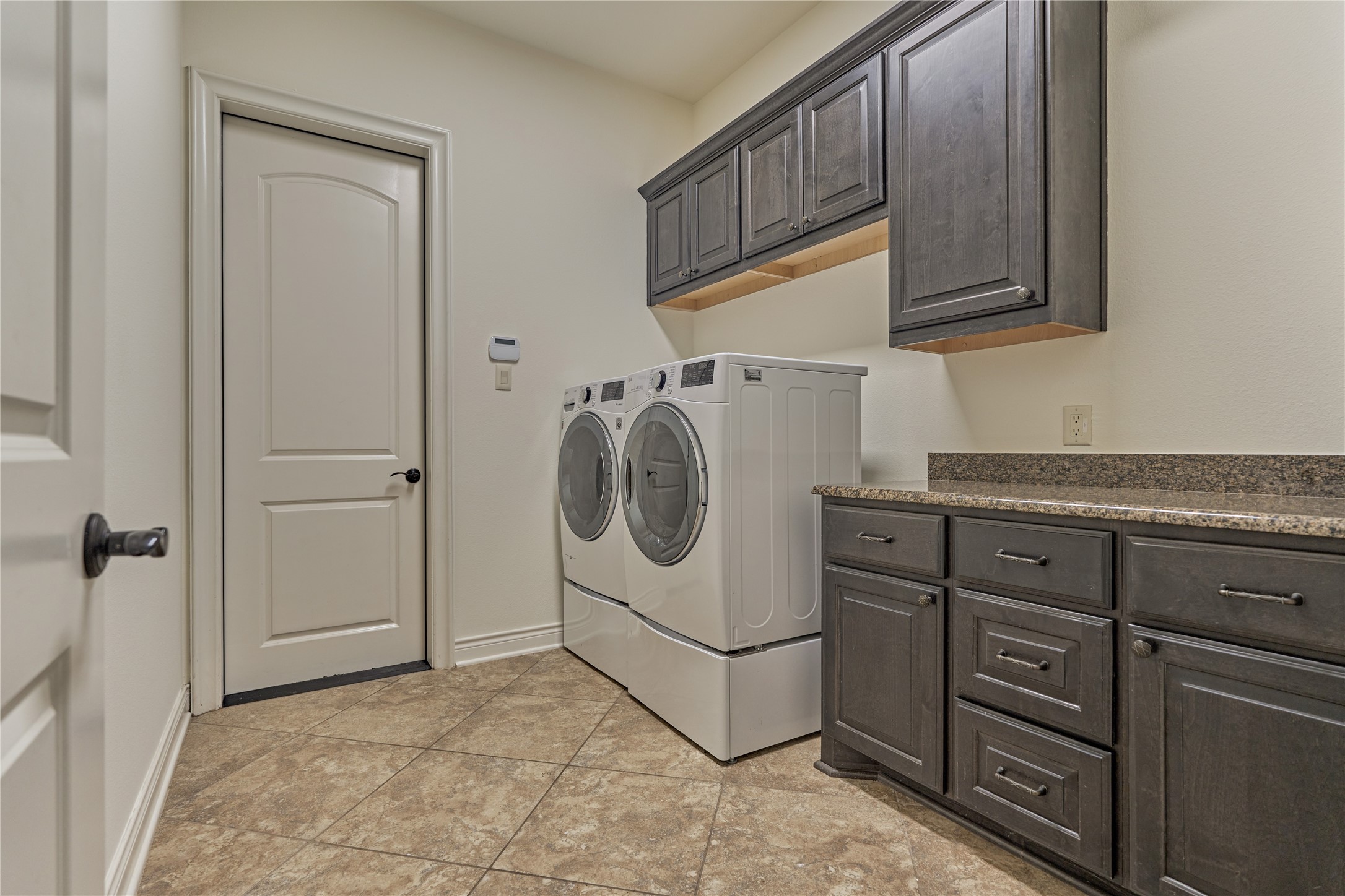 108 Evergreen Oak Drive Conroe, TX 77384 - Photo 28 of 45 This photo showcases a spacious laundry room featuring a modern washer and dryer set. The room has ample storage with dark wood cabinets above and below a granite countertop, and it has tiled flooring for easy maintenance.