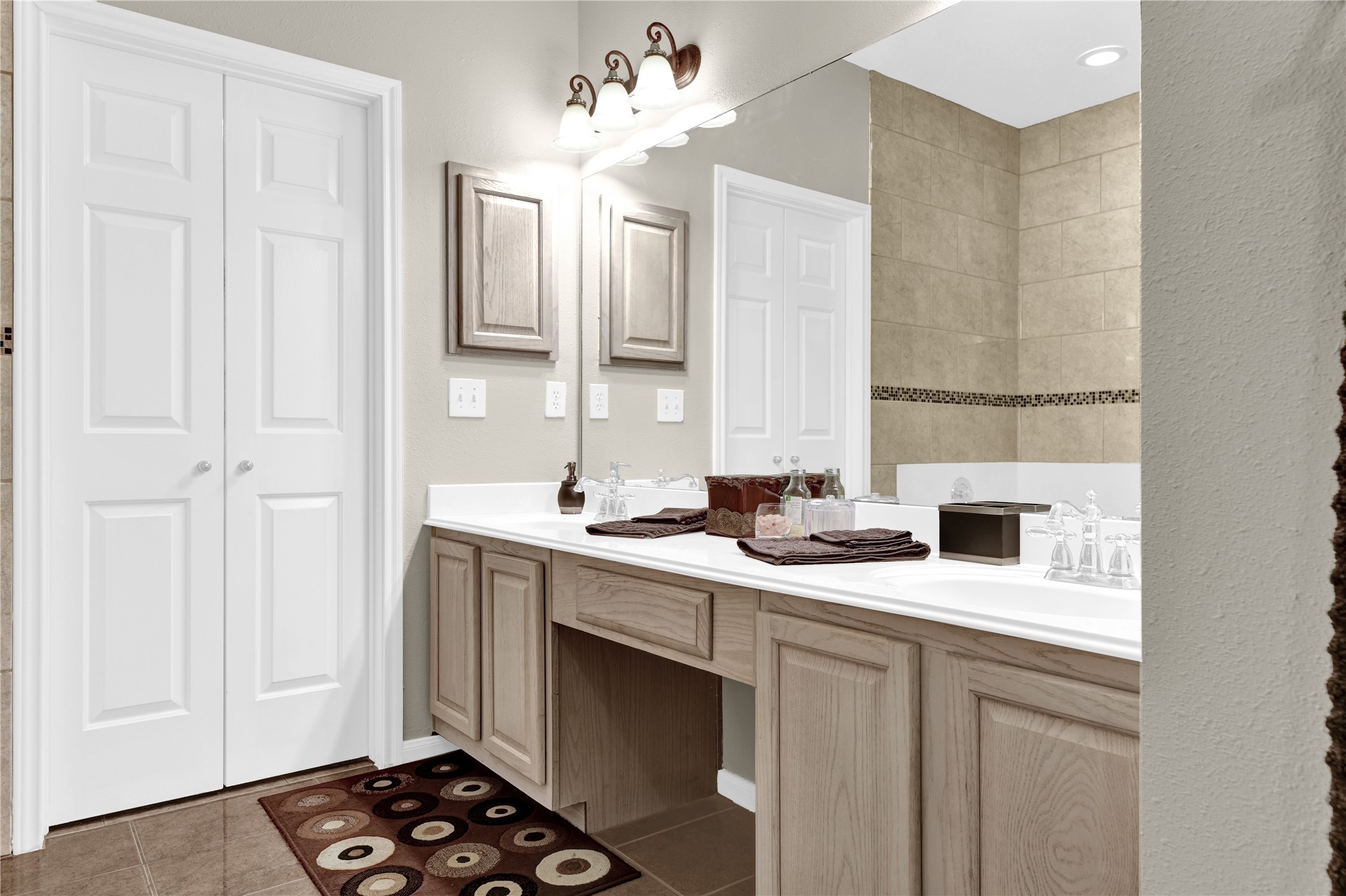 14420 Walters Road, Unit 16 Houston, TX 77014 - Photo 14 of 27 This bathroom features a double vanity with light wood cabinets and a large mirror. It has recessed lighting and stylish fixtures, complemented by a neutral tile backdrop and a decorative border in the shower area. The space offers a clean and modern look with ample storage.