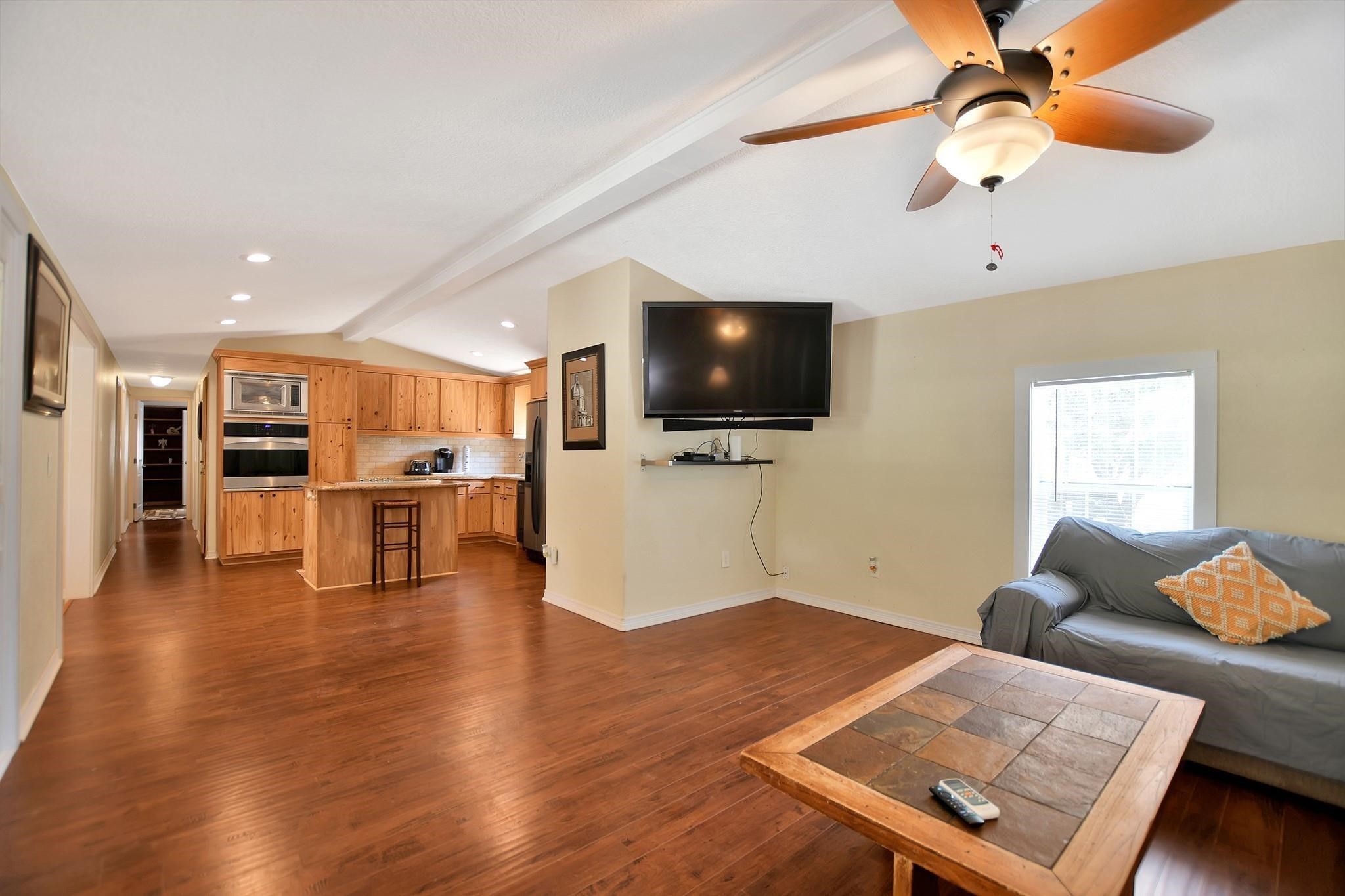 29115 Cherokee Lane Waller, TX 77484 - Photo 11 of 50 This spacious open-concept living area features hardwood floors, a cozy corner kitchen with wooden cabinetry, and a ceiling fan. The room is well-lit with natural light from a large window, and there's a mounted TV for entertainment.