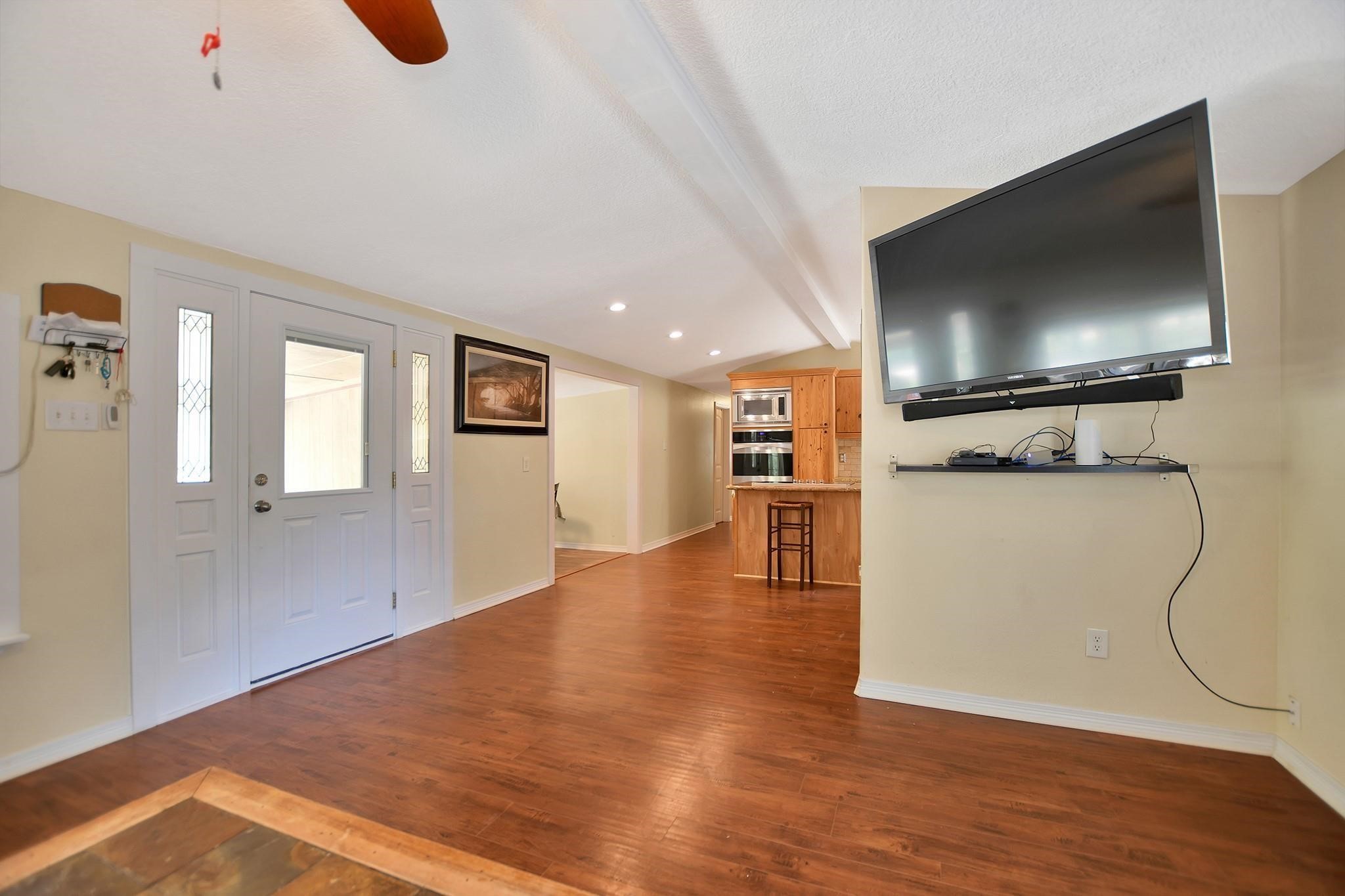 29115 Cherokee Lane Waller, TX 77484 - Photo 13 of 50 Spacious living area with hardwood floors, featuring a wall-mounted TV and a modern open layout. The room includes a stylish kitchen with built-in appliances, a breakfast bar, and ample natural light through a glass-paneled front door. Ideal for entertaining and comfortable living.