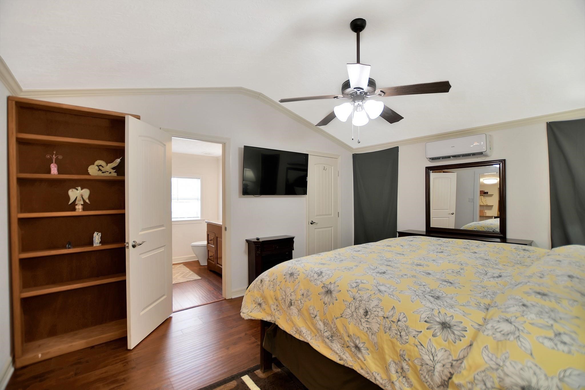 29115 Cherokee Lane Waller, TX 77484 - Photo 23 of 50 Cozy bedroom with vaulted ceiling and wood flooring. Features a ceiling fan, wall-mounted TV, and open shelving. The room includes an ensuite bathroom and a mirrored dresser under an air conditioning unit.