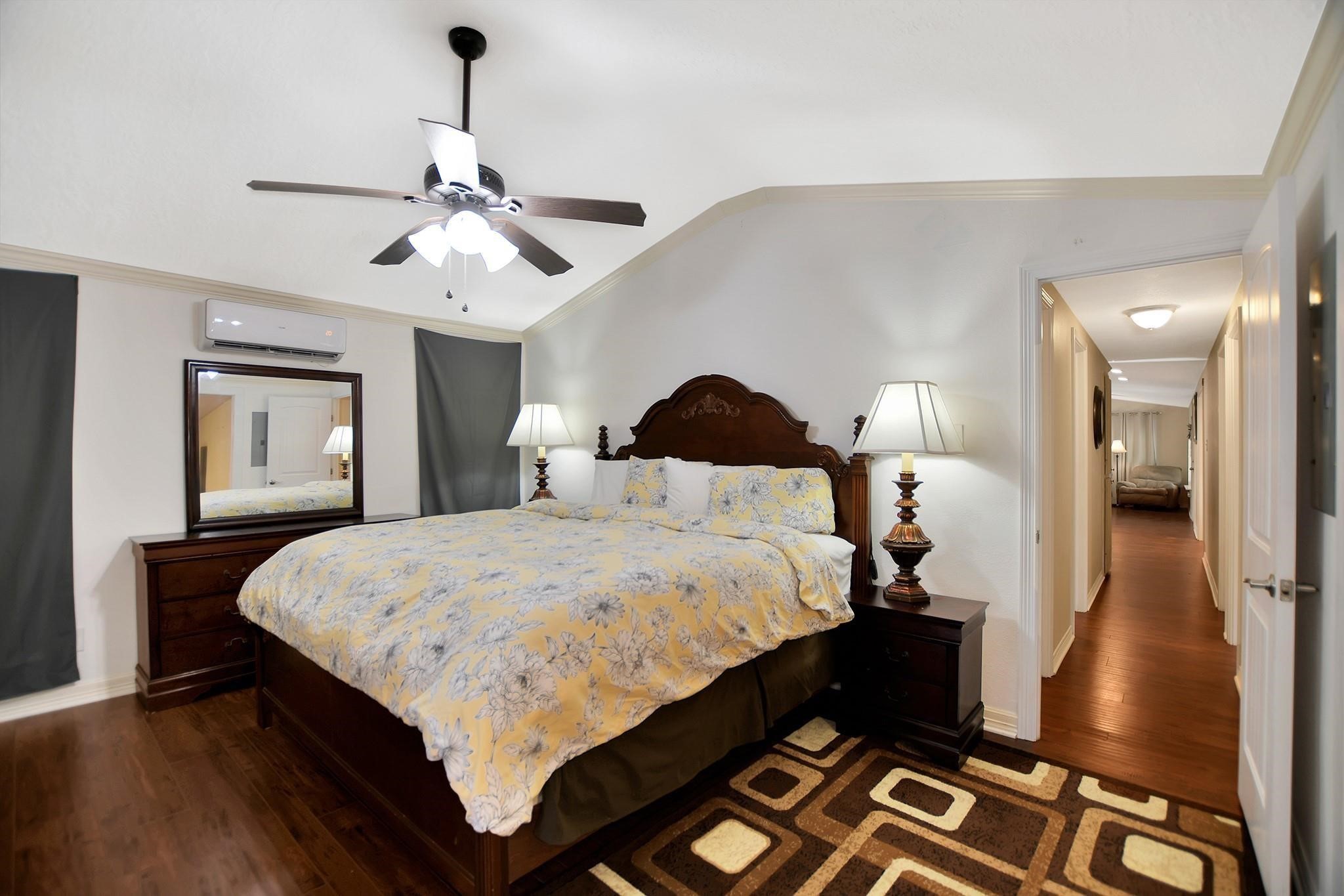29115 Cherokee Lane Waller, TX 77484 - Photo 24 of 50 This bedroom features a large bed with an ornate headboard, wooden nightstands with lamps, and a ceiling fan. It has hardwood flooring and a patterned rug, with a view down a hallway that leads to another room.