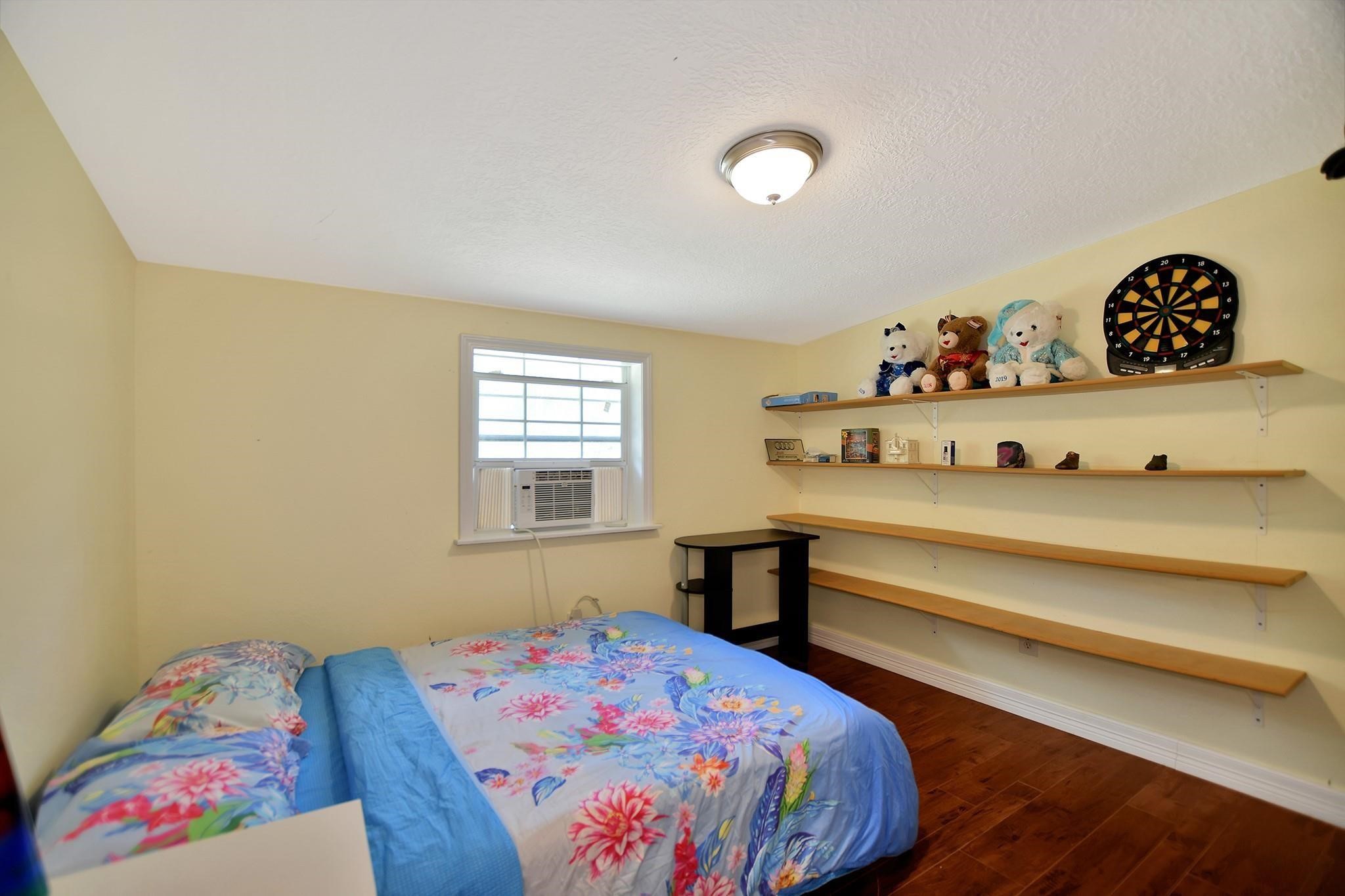 29115 Cherokee Lane Waller, TX 77484 - Photo 31 of 50 Cozy bedroom with a floral bedspread, window AC unit, and light yellow walls. Features wooden shelves with decor and a dartboard, plus warm wooden flooring. Ideal for a child's room or guest space.