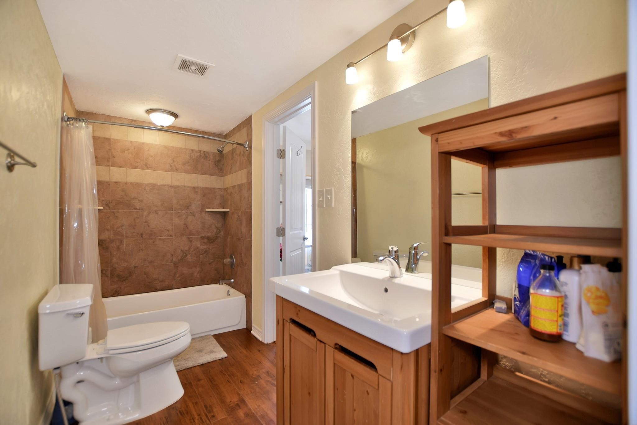 29115 Cherokee Lane Waller, TX 77484 - Photo 33 of 50 This bathroom features a cozy design with a tiled shower-bathtub combo, a wooden vanity with a sink, a large mirror, and additional shelving. The warm lighting and wood flooring add to the inviting atmosphere.