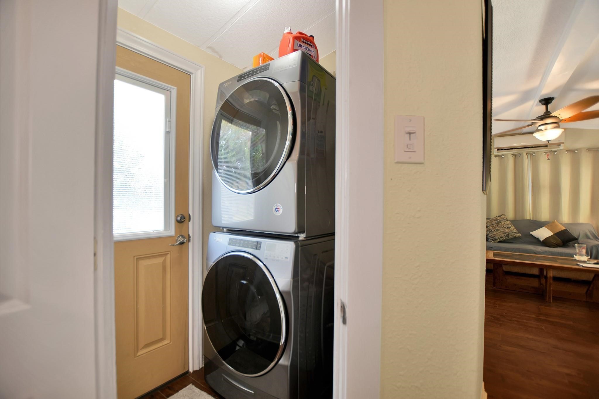 29115 Cherokee Lane Waller, TX 77484 - Photo 34 of 50 This photo shows a compact laundry area with a stacked washer and dryer next to a door with frosted glass. Adjacent is a cozy living room with a ceiling fan, wooden flooring, and a seating area with cushions.
