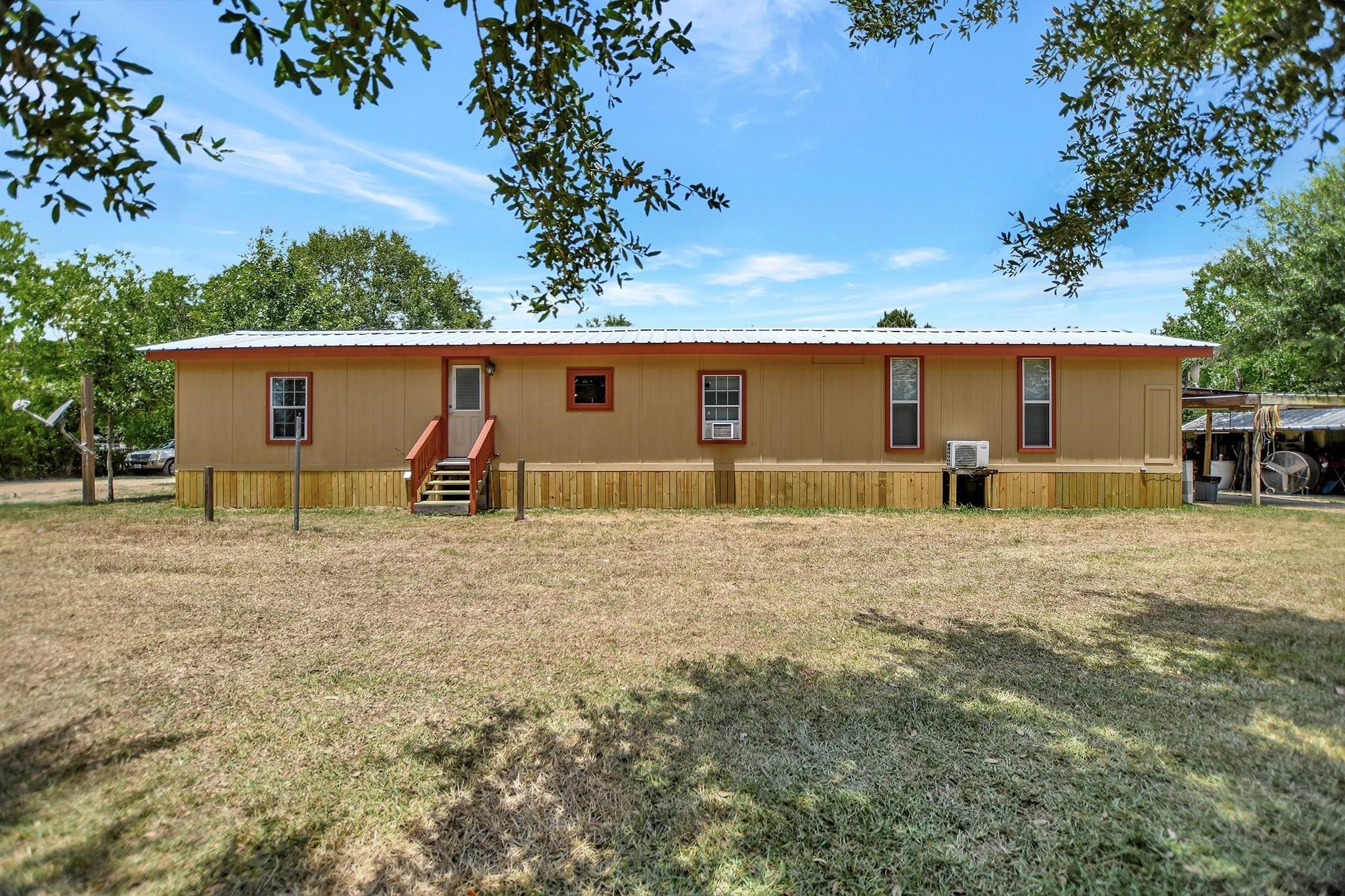 29115 Cherokee Lane Waller, TX 77484 - Photo 36 of 50 This photo shows a single-story, tan mobile home with a red trim and a metal roof. It features a small wooden porch with stairs leading to the entrance. The home is surrounded by an expansive grassy yard with trees, offering a peaceful, rural setting.