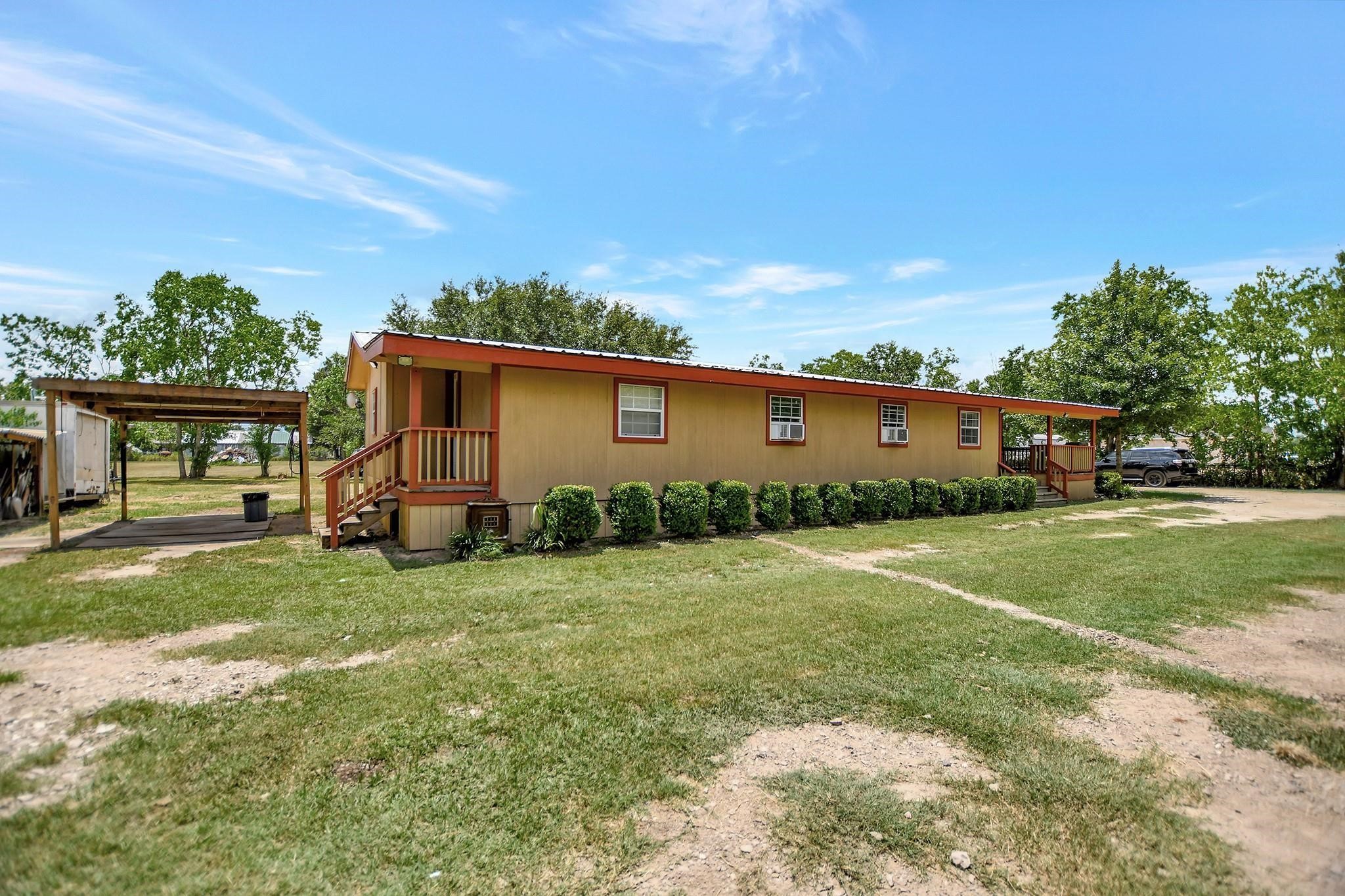 29115 Cherokee Lane Waller, TX 77484 - Photo 38 of 50 This photo shows a single-story mobile home with a warm exterior, featuring a small porch and a covered carport. It is surrounded by a spacious yard with some greenery and mature trees, set against a clear blue sky.