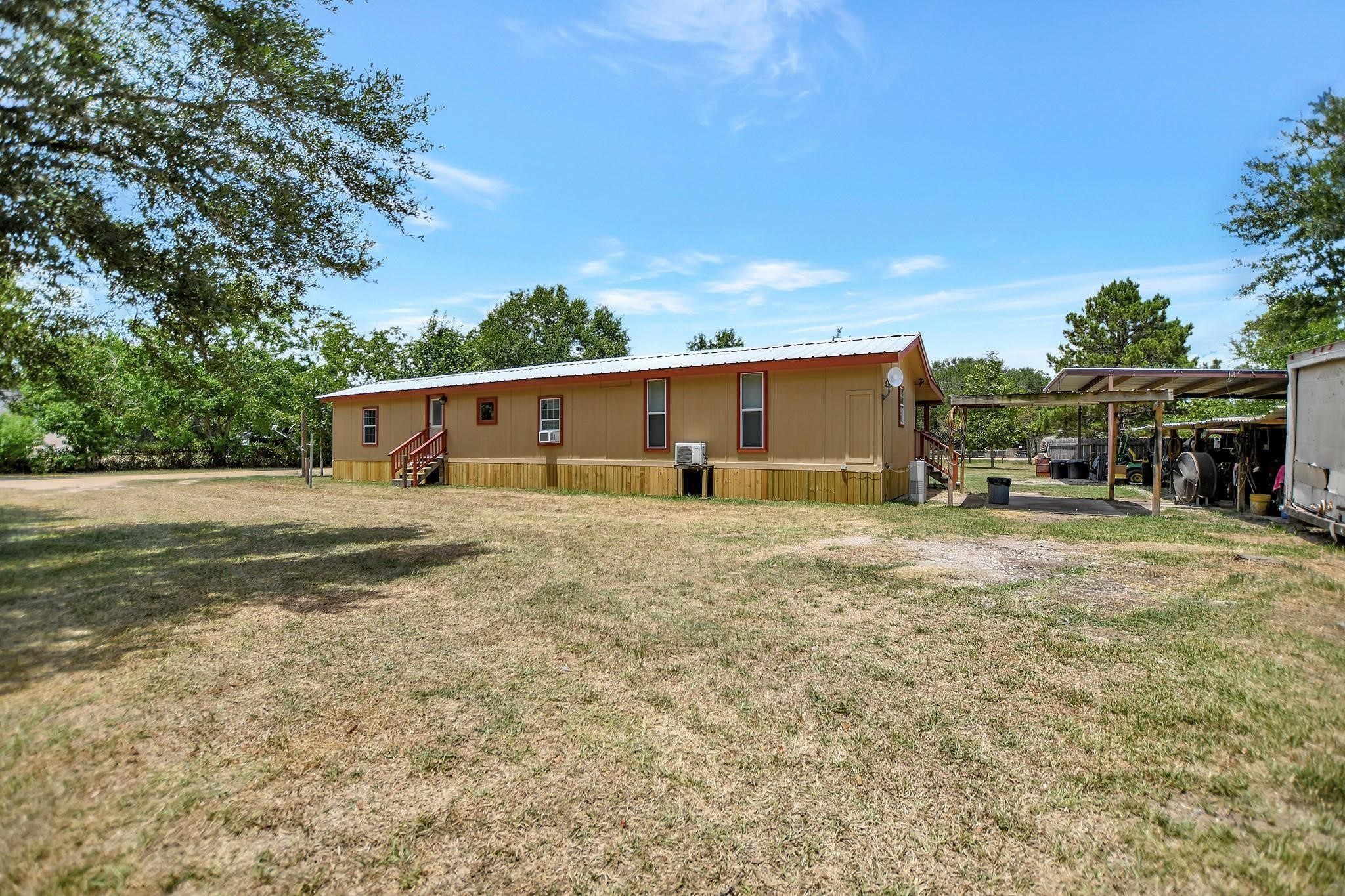 29115 Cherokee Lane Waller, TX 77484 - Photo 40 of 50 This photo showcases a single-story mobile home with a metal roof, situated on a spacious, grassy lot. It features a covered carport and mature trees, offering ample outdoor space and shade.