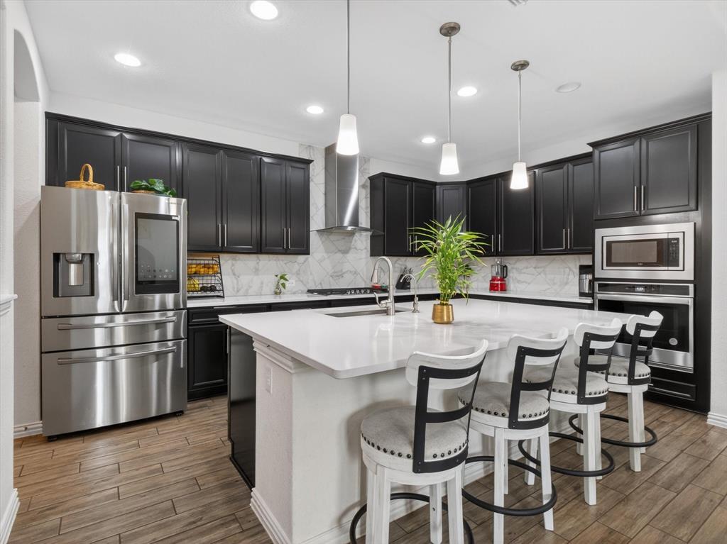 636 Corner Post Path Celina, TX 75009 - Photo 13 of 40 Modern kitchen featuring black cabinetry, a spacious island with seating, stainless steel appliances, marble-look backsplash, and wood-finish flooring
