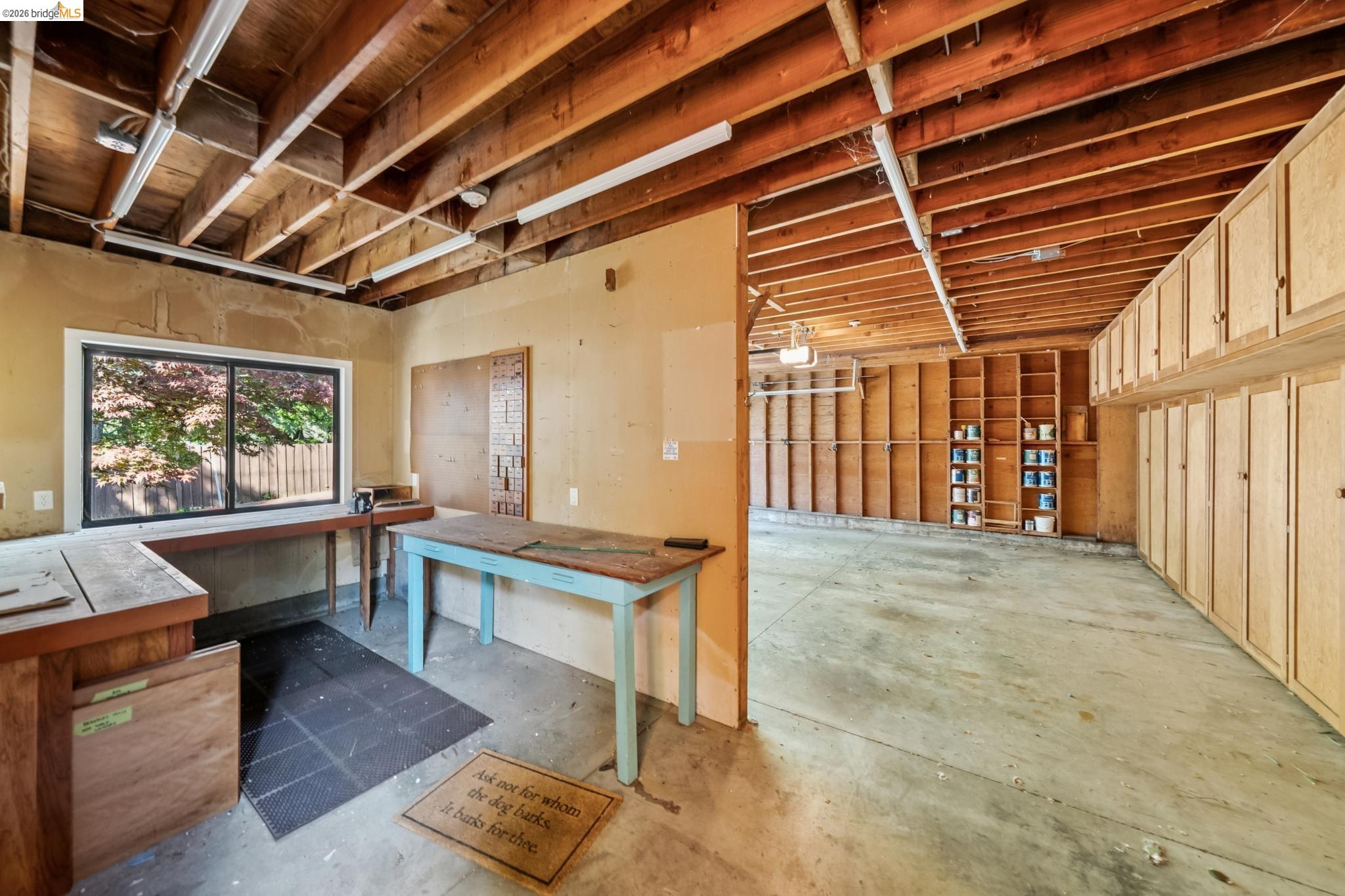 576 Vistamont Avenue Berkeley, CA 94708 - Photo 33 of 33 Spacious workshop and garage area featuring extensive built-in cabinetry, exposed wood ceiling beams, concrete flooring, a large window, and fluorescent overhead lighting