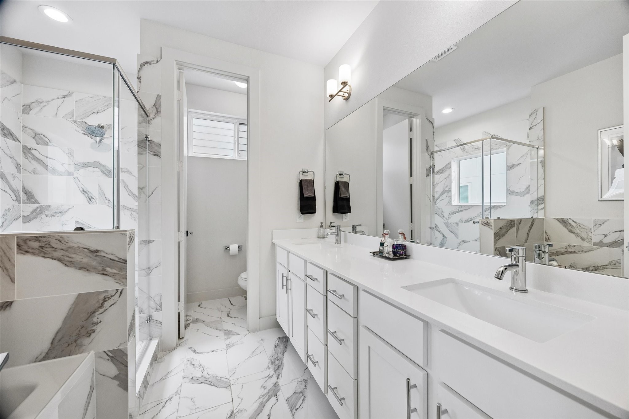 1310 Hussion Street Houston, TX 77003 - Photo 19 of 34 This bathroom features a sleek, modern design with white cabinetry, a long double vanity, and elegant marble-patterned tile throughout. It includes a spacious glass-enclosed shower and a separate toilet area, offering both style and functionality.