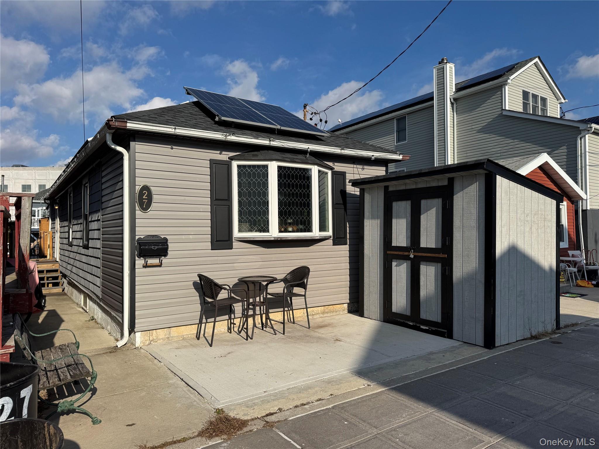 27 West 11th Road Queens, NY 11693 - Photo 2 of 18 Front of house with roof mounted solar panels, a storage unit, a patio area, and roof with shingles