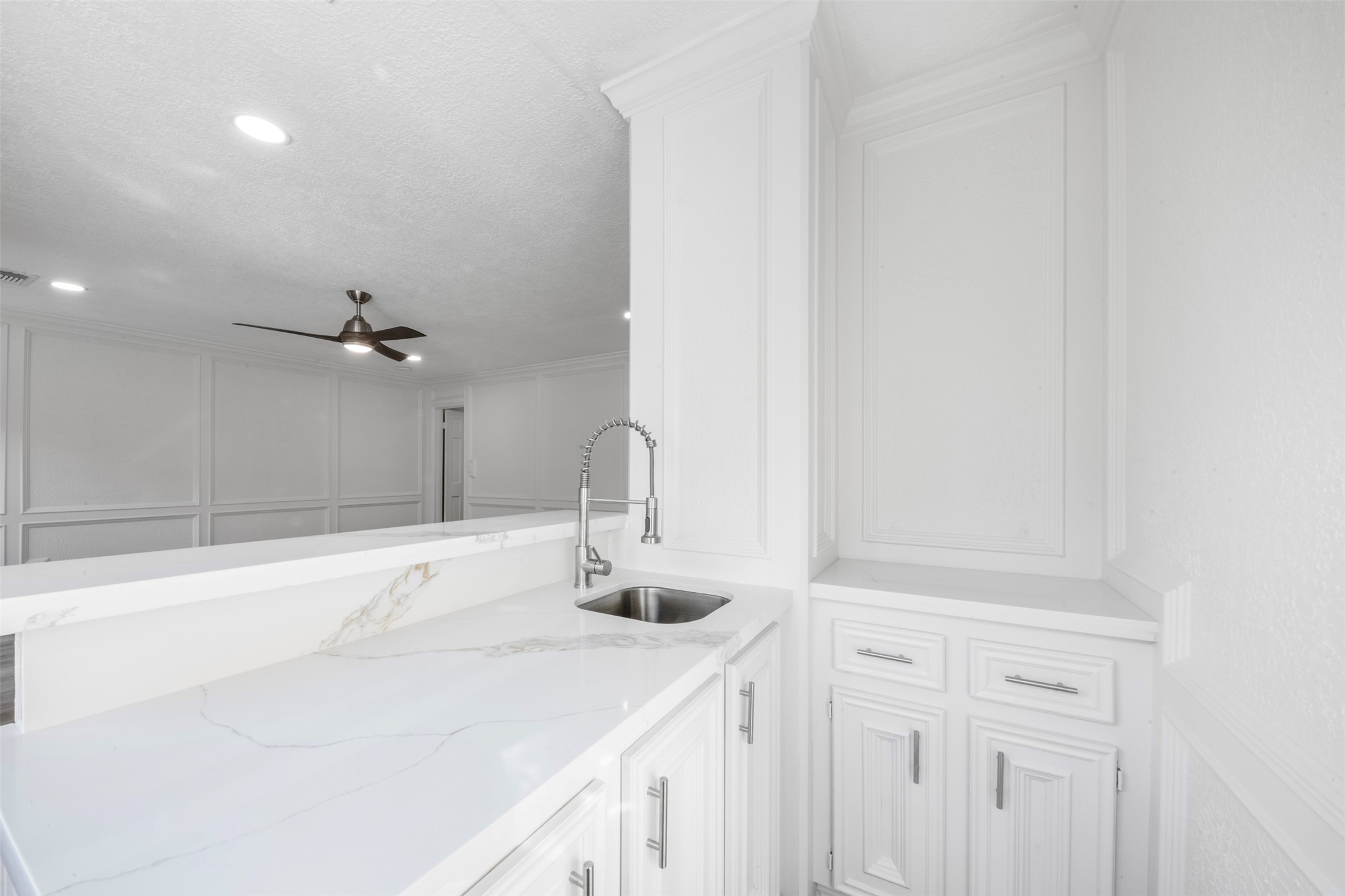 9543 Enstone Circle Spring, TX 77379 - Photo 32 of 50 This photo showcases a bright, modern kitchen with white cabinetry and a sleek marble countertop. It features a stylish gooseneck faucet and opens to a spacious living area with a ceiling fan and recessed lighting.