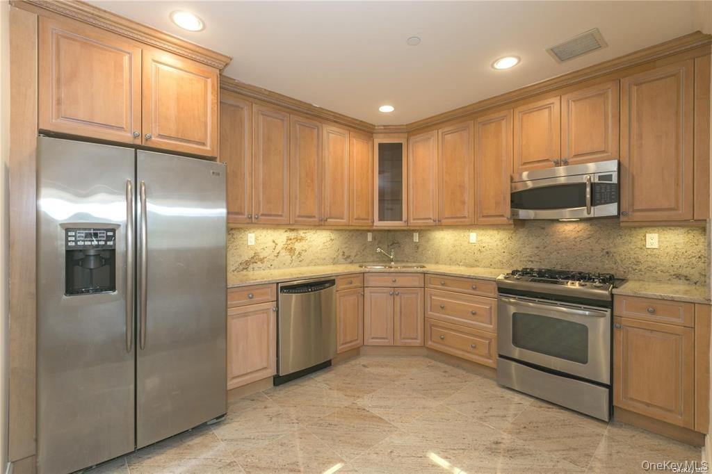 171 Great Neck Road, Unit 1N Great Neck, NY 11021 - Photo 9 of 15 Kitchen featuring stainless steel appliances, light stone counters, glass insert cabinets, decorative backsplash, and brown cabinets