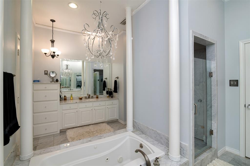 5920 Encore Drive Dallas, TX 75240 - Photo 20 of 34 Contemporary bathroom featuring a jetted tub set in a marble surround, a dual vanity with white cabinetry and light-toned countertops, a glass-enclosed shower with marble-look tile, and decorative chandeliers