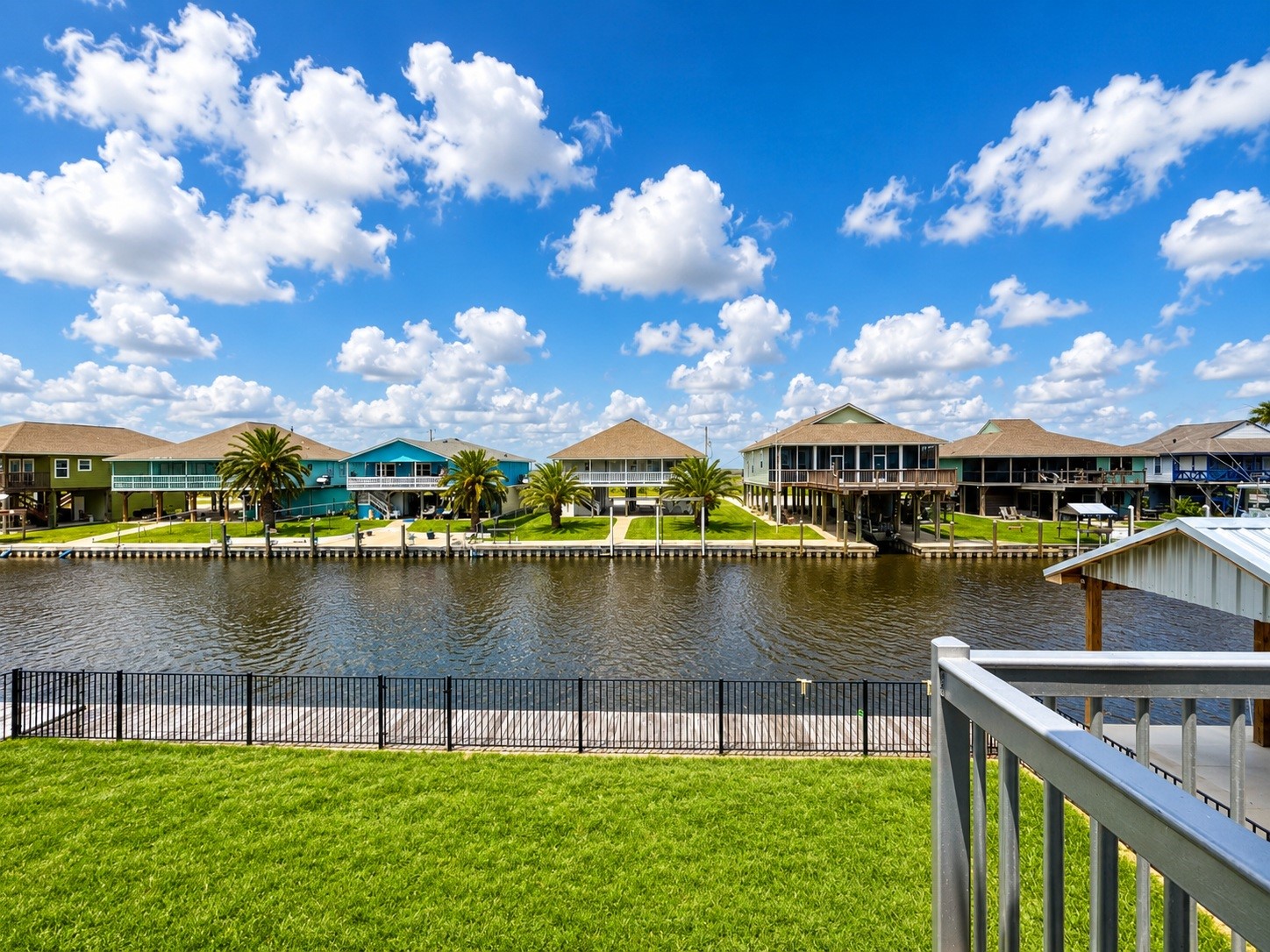 135 Sand Shoals Road Freeport, TX 77541 - Photo 3 of 20 This photo showcases a picturesque waterfront view with charming houses on stilts across a serene canal. A lush green lawn is in the foreground, bordered by a metal fence, offering a peaceful and scenic environment. The clear blue sky dotted with fluffy clouds enhances the tranquil setting.