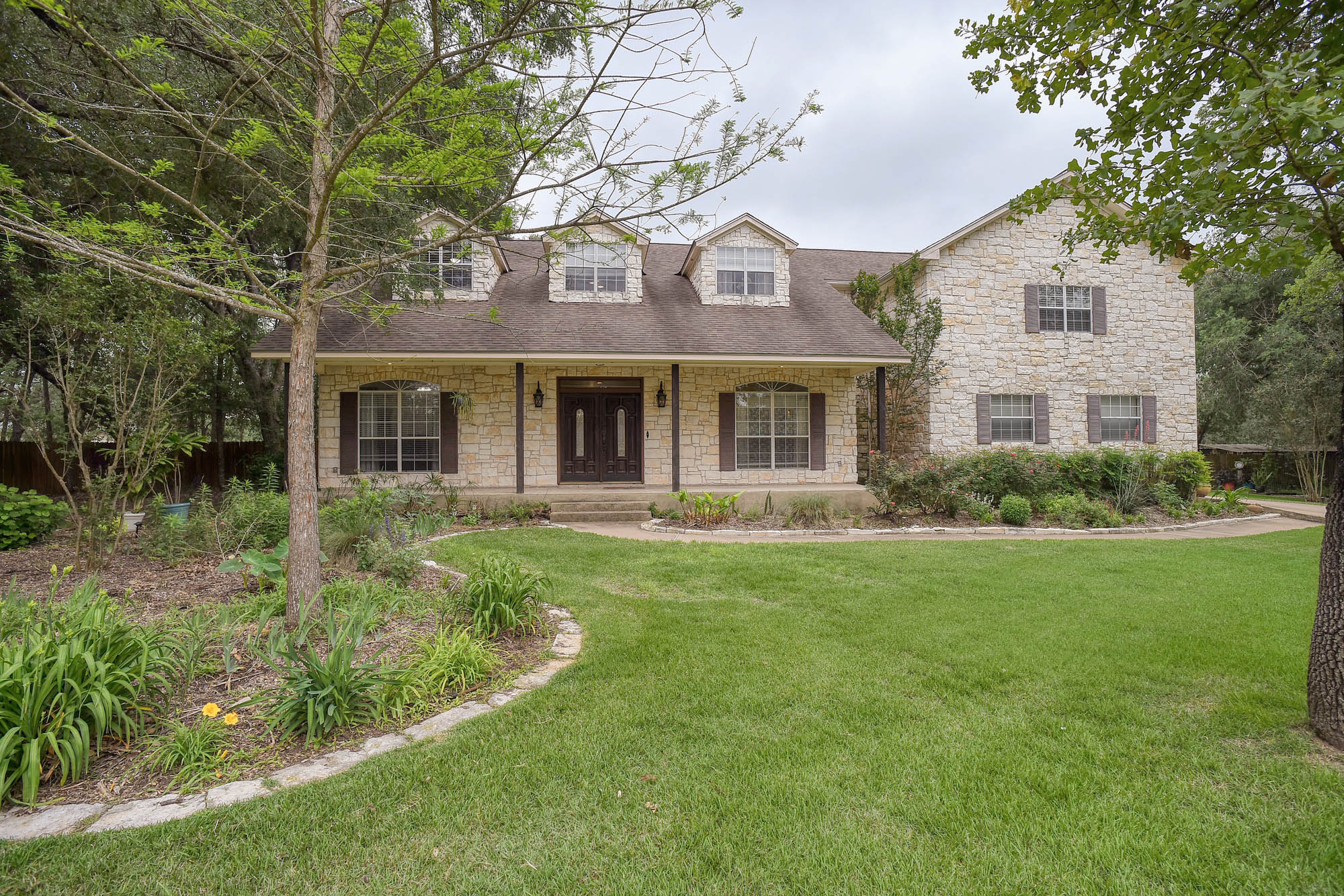 Stone-clad exterior featuring a covered front porch with columns, dark double entry doors, dormer windows, and dark louvered shutters