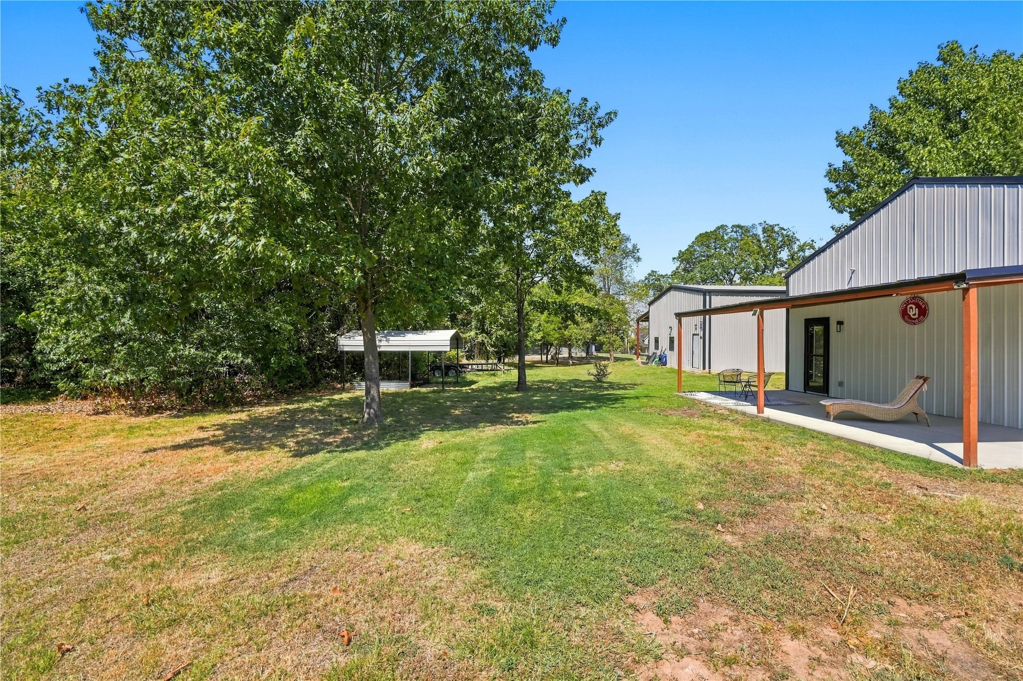Lawn area with mature tree, a covered patio, and structures featuring metal siding and covered entryways