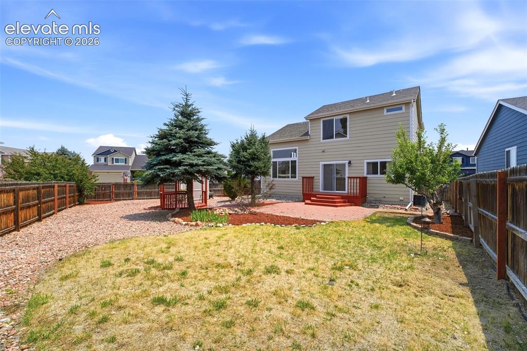 7904 Coffee Road Peyton, CO 80831 - Photo 35 of 48 Spacious backyard featuring a tan exterior home with a red-stained deck, mature evergreen trees, and a wooden fence